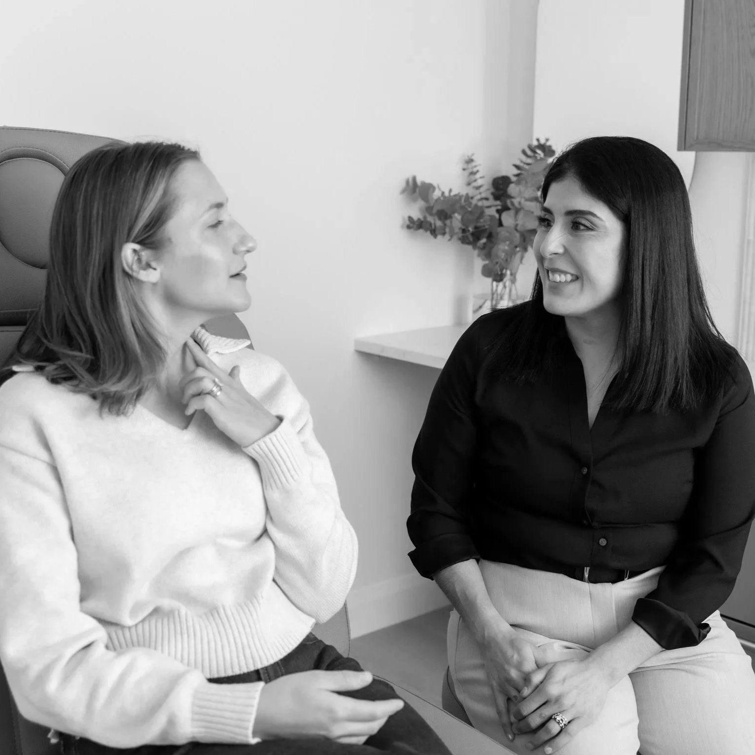 Two women sitting close, engaging in a conversation with smiling expressions, one touching her neck and the other with hand on her lap, in a room with a small table and flowers in the background.