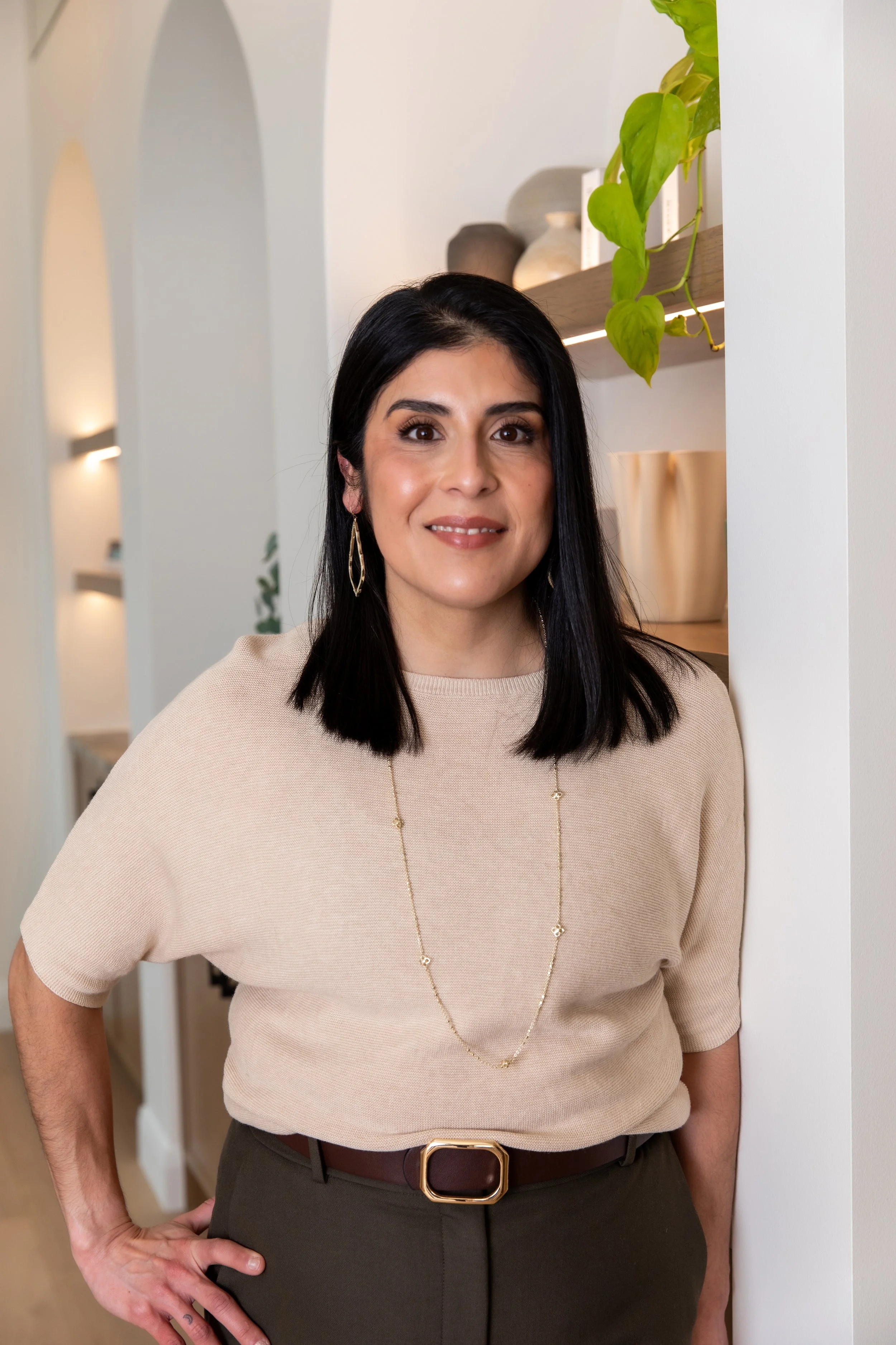 A woman with shoulder-length black hair, wearing a beige short-sleeve sweater, dark pants, and gold jewelry, standing in a modern, white-walled interior.