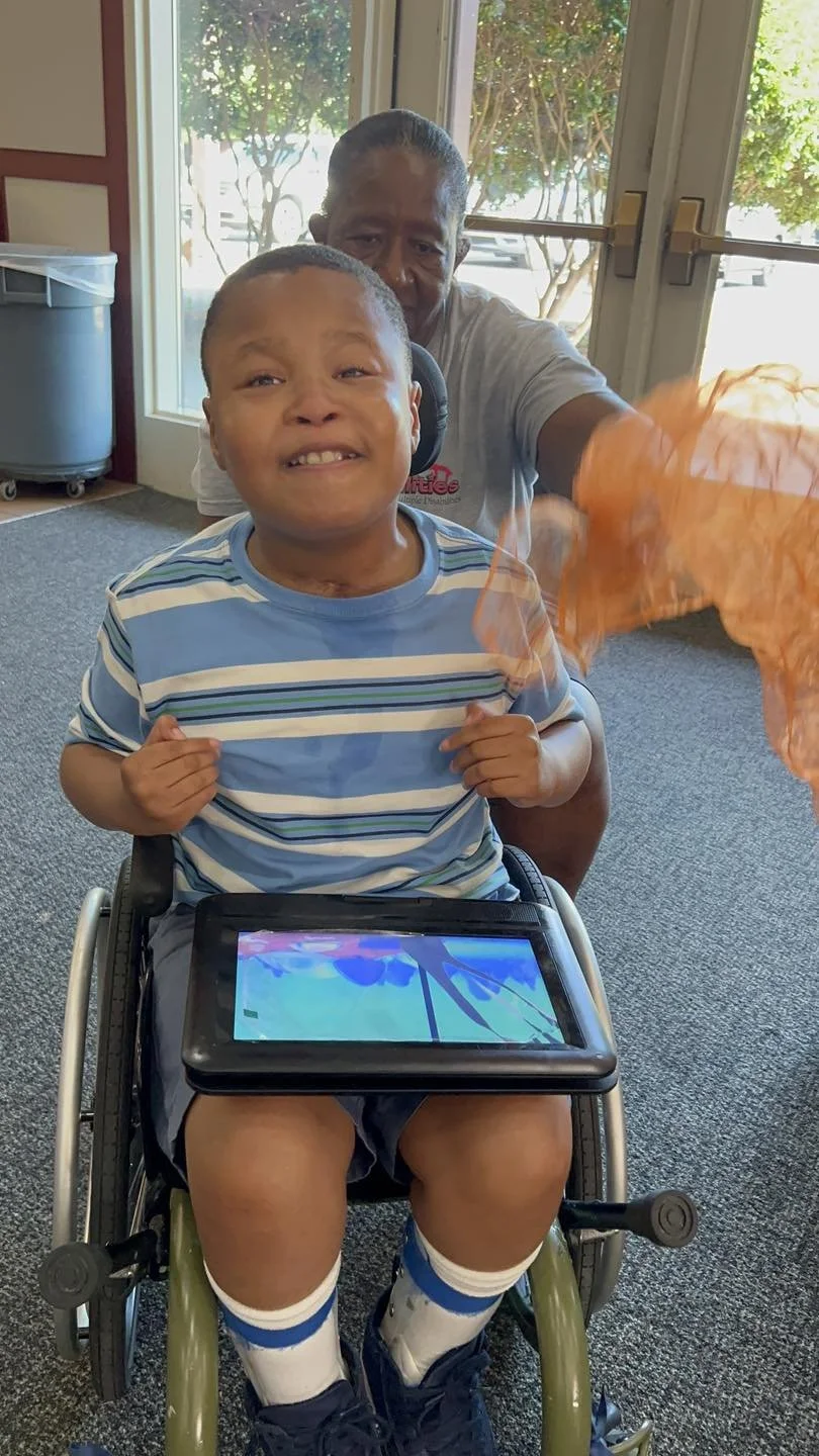 A young boy in a wheelchair with a tablet on his lap, smiling and pulling at his shirt, and an older man behind him smiling, holding an orange tissue or cloth, inside a room with large windows and a view of trees outside.