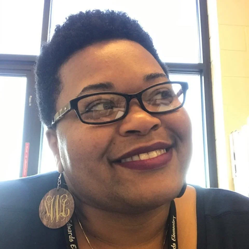 A smiling woman with short curly hair wearing glasses, a large earring, and a lanyard in front of a window.