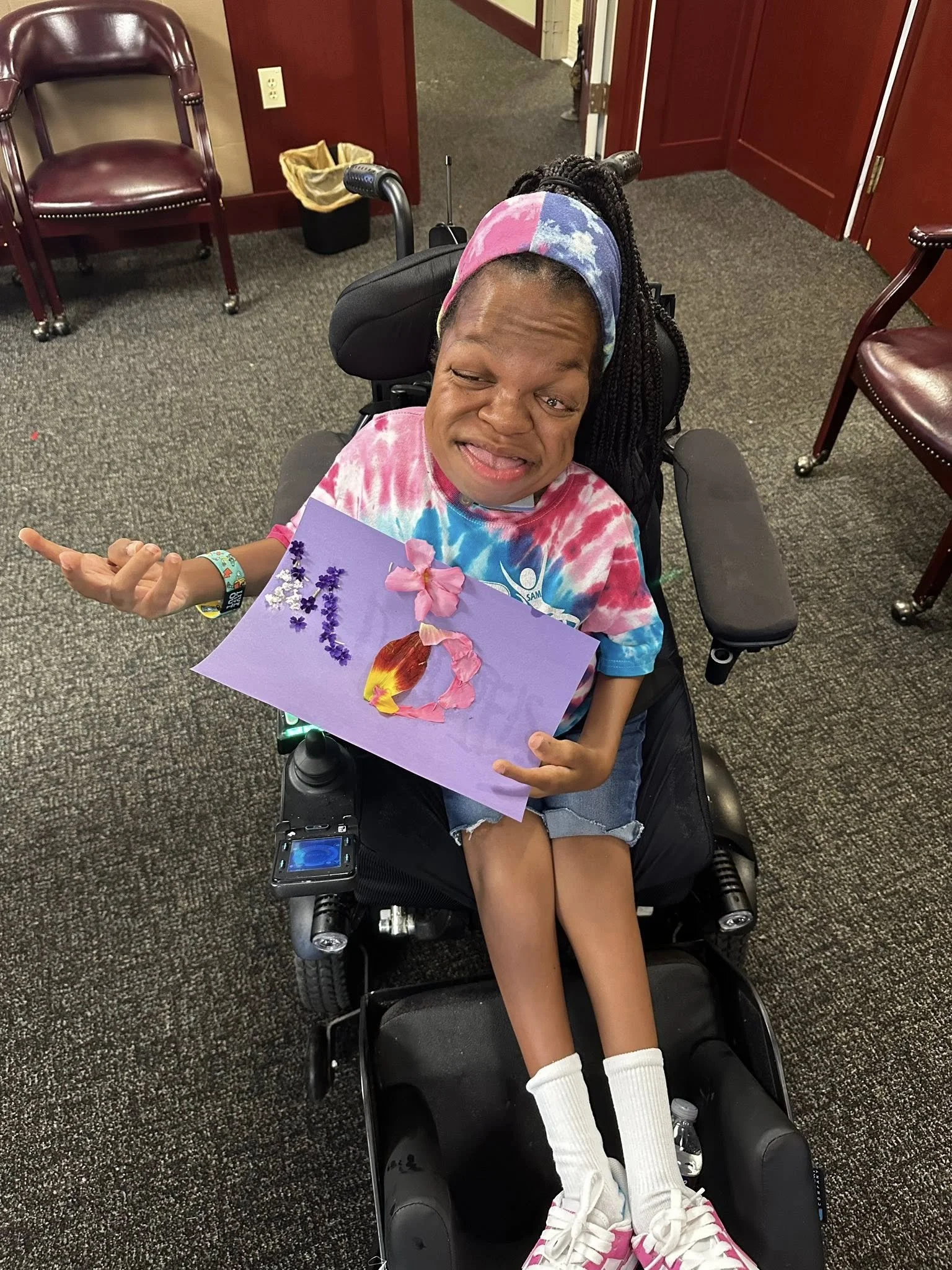 A young girl sitting in a motorized wheelchair, wearing a tie-dye shirt, shorts, white socks, and pink sneakers. She is holding a handmade card decorated with flowers and a fire image, and is making a playful face.