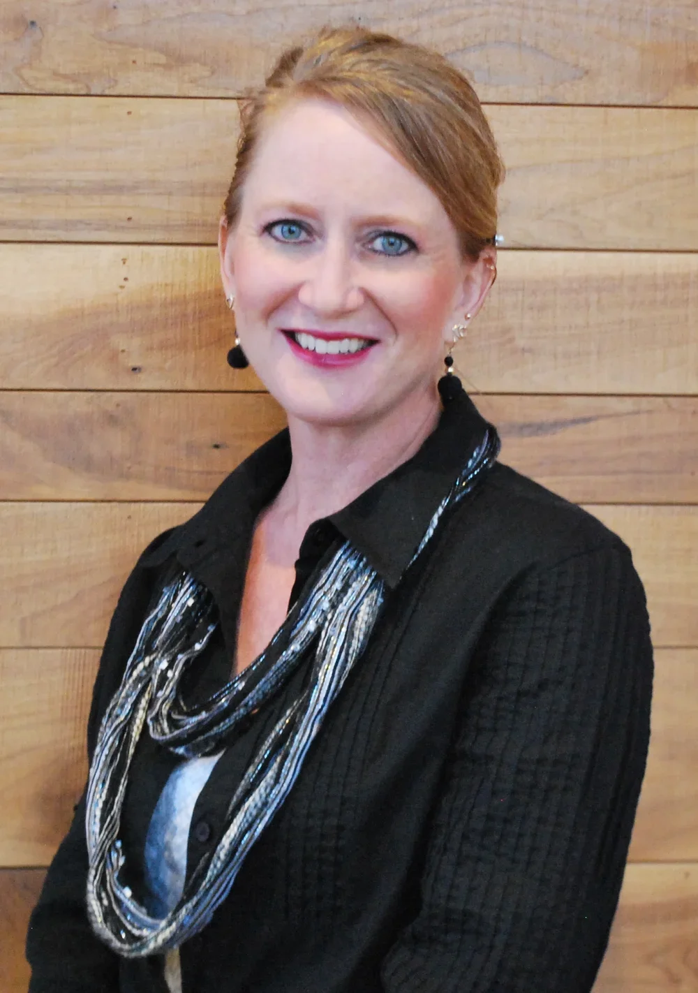A woman with short red hair, blue eyes, and fair skin smiling, wearing a black top with a patterned scarf and earrings, standing in front of a wooden wall.