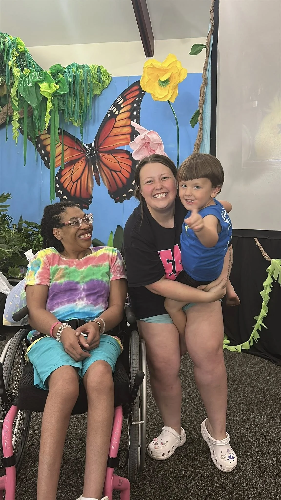 Two girls, one in a wheelchair and one sitting on an adult's lap, smiling and enjoying a celebration or event with a colorful butterfly and flower backdrop.