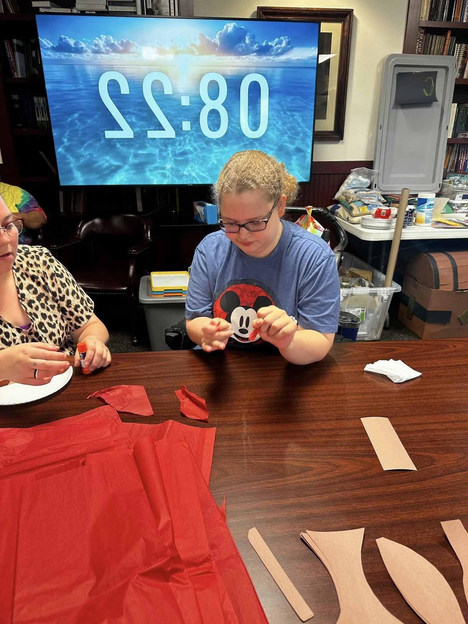 People crafting paper decorations at a table, with a large screen showing a swimming pool scene and the time 08:22.