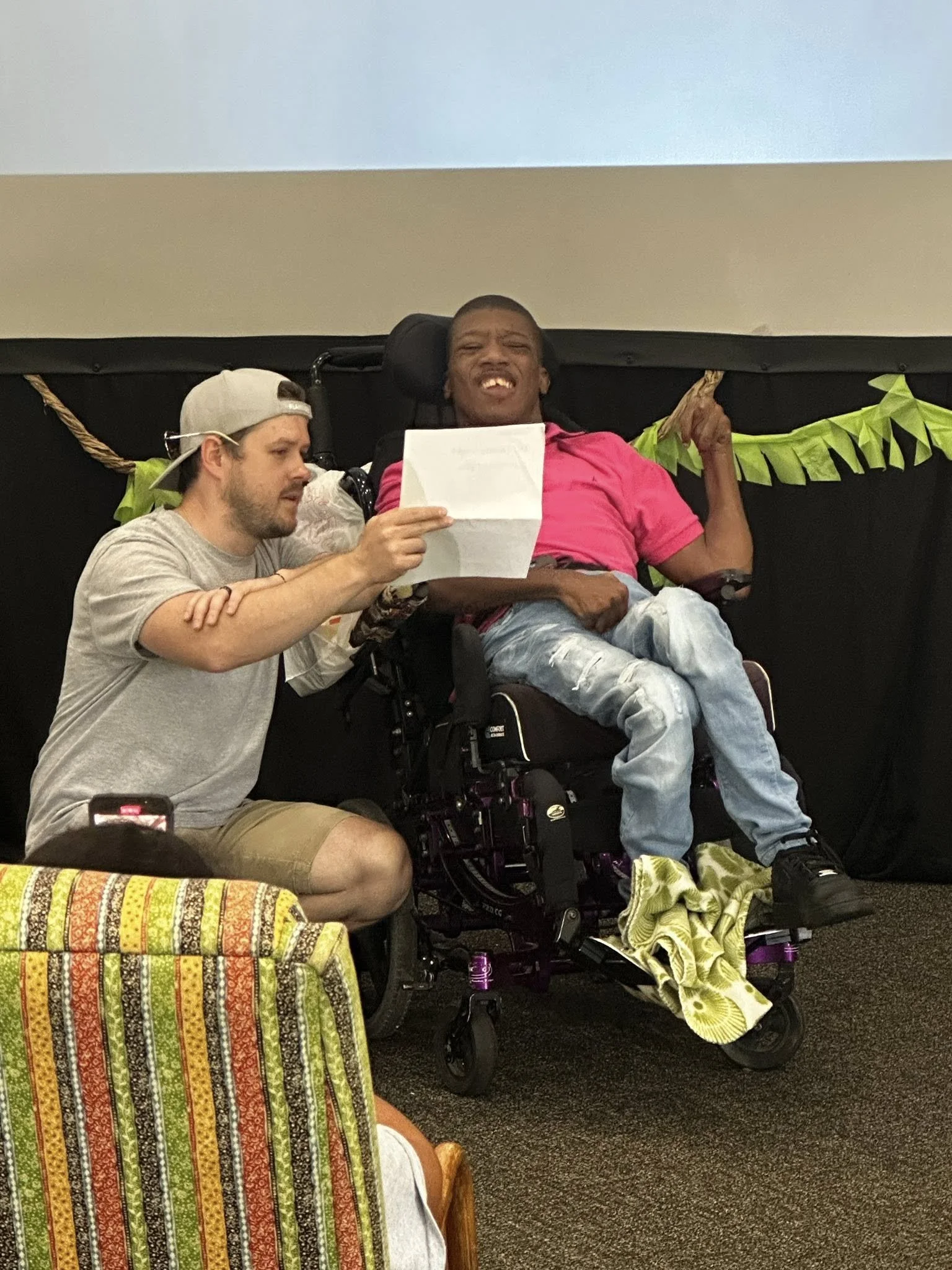 A young man in a pink shirt sitting in a motorized wheelchair, holding a piece of paper, smiling and pointing upward with one finger, while a young man kneeling beside him looks at the paper. There are party decorations behind them and a group of people sitting nearby.