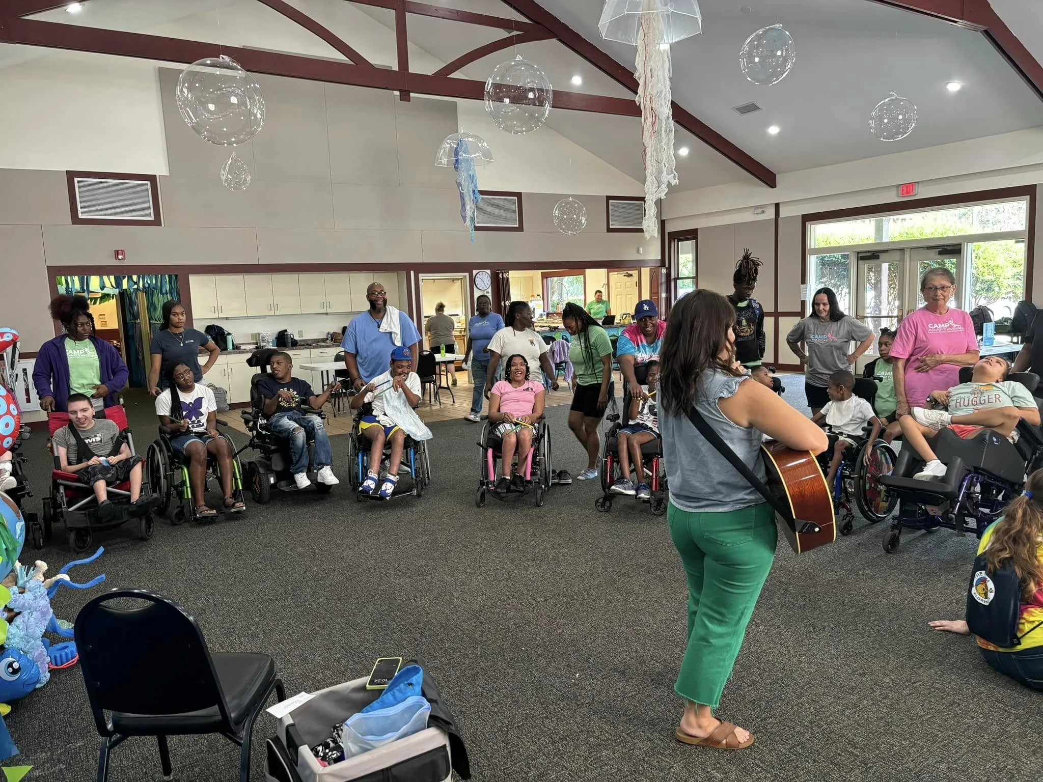 A woman with dark hair playing guitar in front of a group of children in wheelchairs in a spacious indoor area with hanging decorations and large windows.