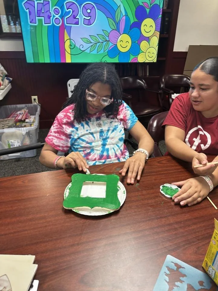 A young girl with glasses and curly hair, wearing a tie-dye t-shirt, decorating a cake with green icing at a table in a room with a colorful rainbow and smiling flower display on a monitor in the background.