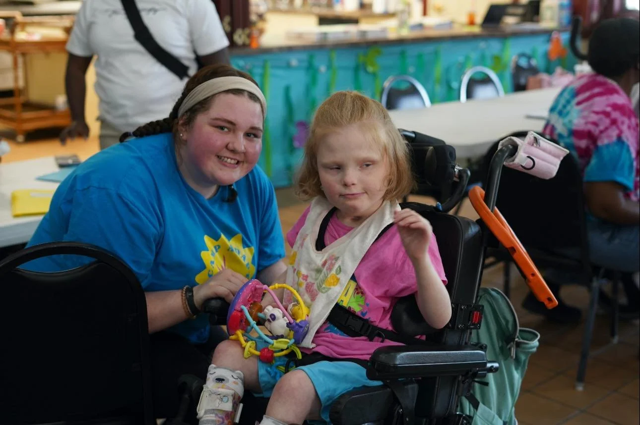 Two girls, one with a developmental disability, sitting together at a table in a community center or classroom. The girl on the left has brown hair, a beige headband, and is smiling. The girl on the right has red hair, wearing a pink shirt and a white bib, with a neutral expression. There are other people and tables in the background.