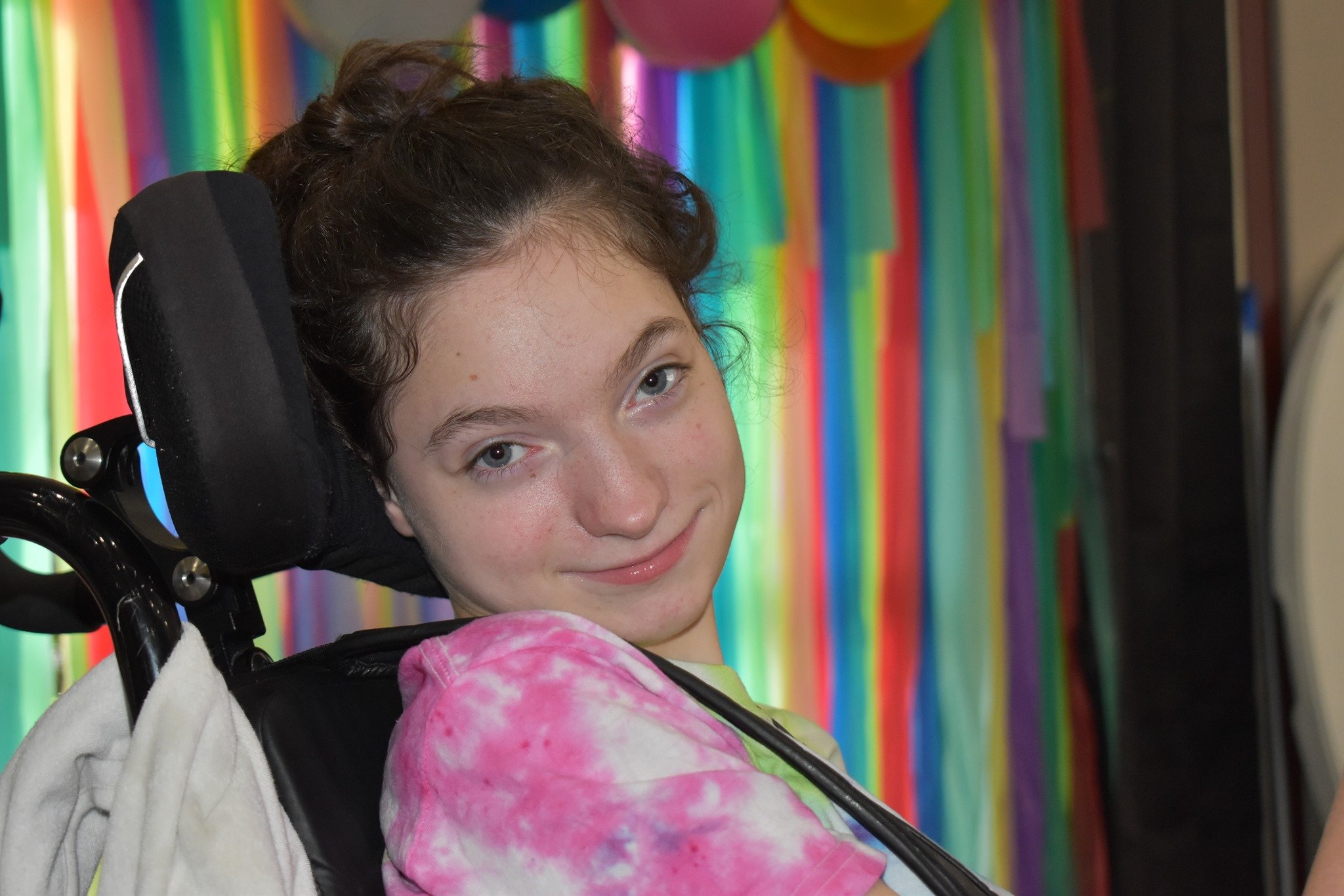 Young girl with cerebral palsy in a wheelchair, wearing a pink tie-dye shirt, smiling at the camera with a colorful rainbow backdrop.