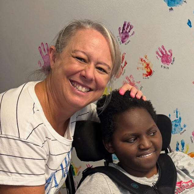 A woman smiling and embracing a young girl in a wheelchair, in front of a wall with colorful handprints and names.