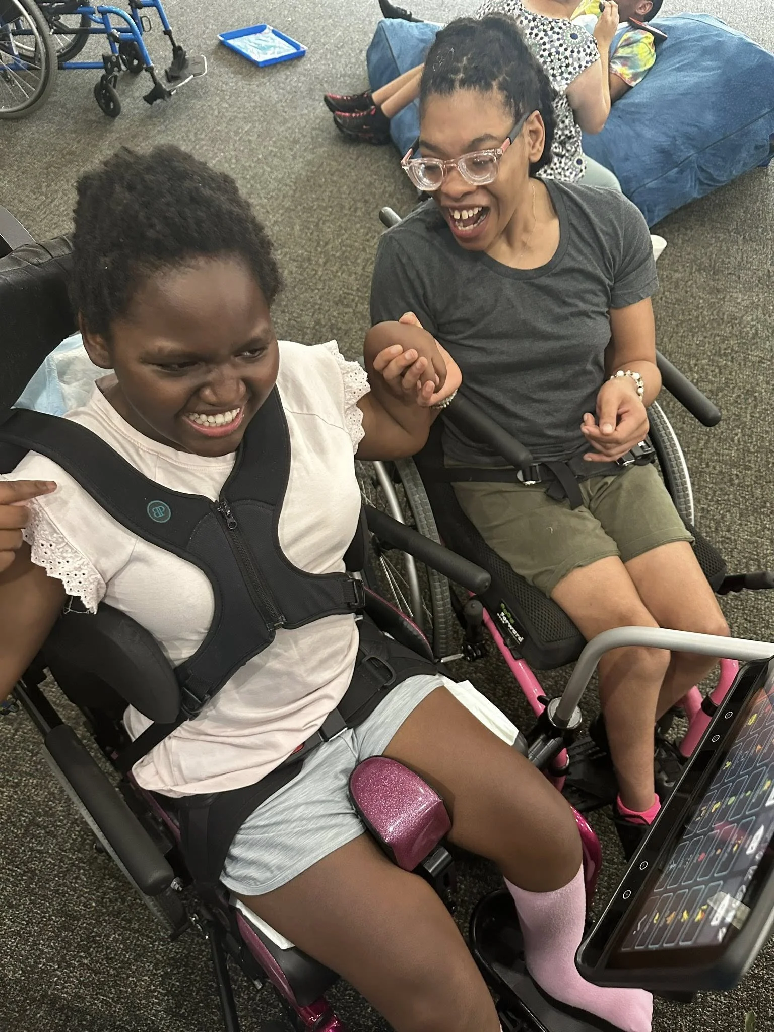 Two women, one in a wheelchair, are holding hands and smiling at each other. The woman in the wheelchair has curly hair, glasses, and is wearing a gray t-shirt and shorts. The other woman has short curly hair, is wearing a white blouse and shorts, and she appears to be happy.