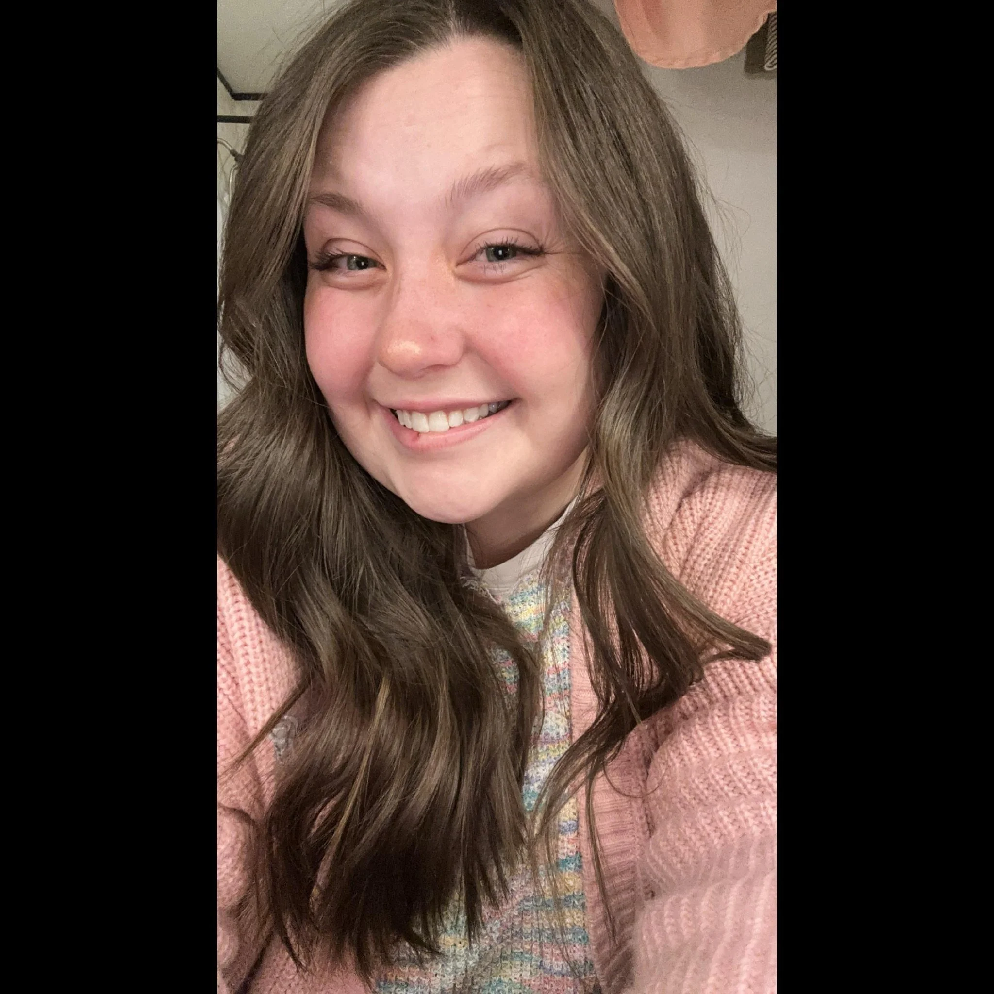 Close-up photo of a young woman with long brown hair, smiling, wearing a pink sweater.