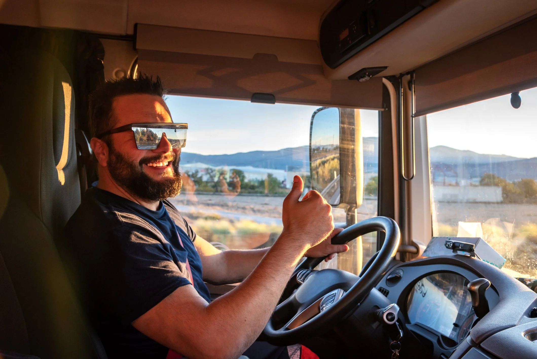 Smiling man driving a truck on a highway with mountains and water in the background, during daytime.