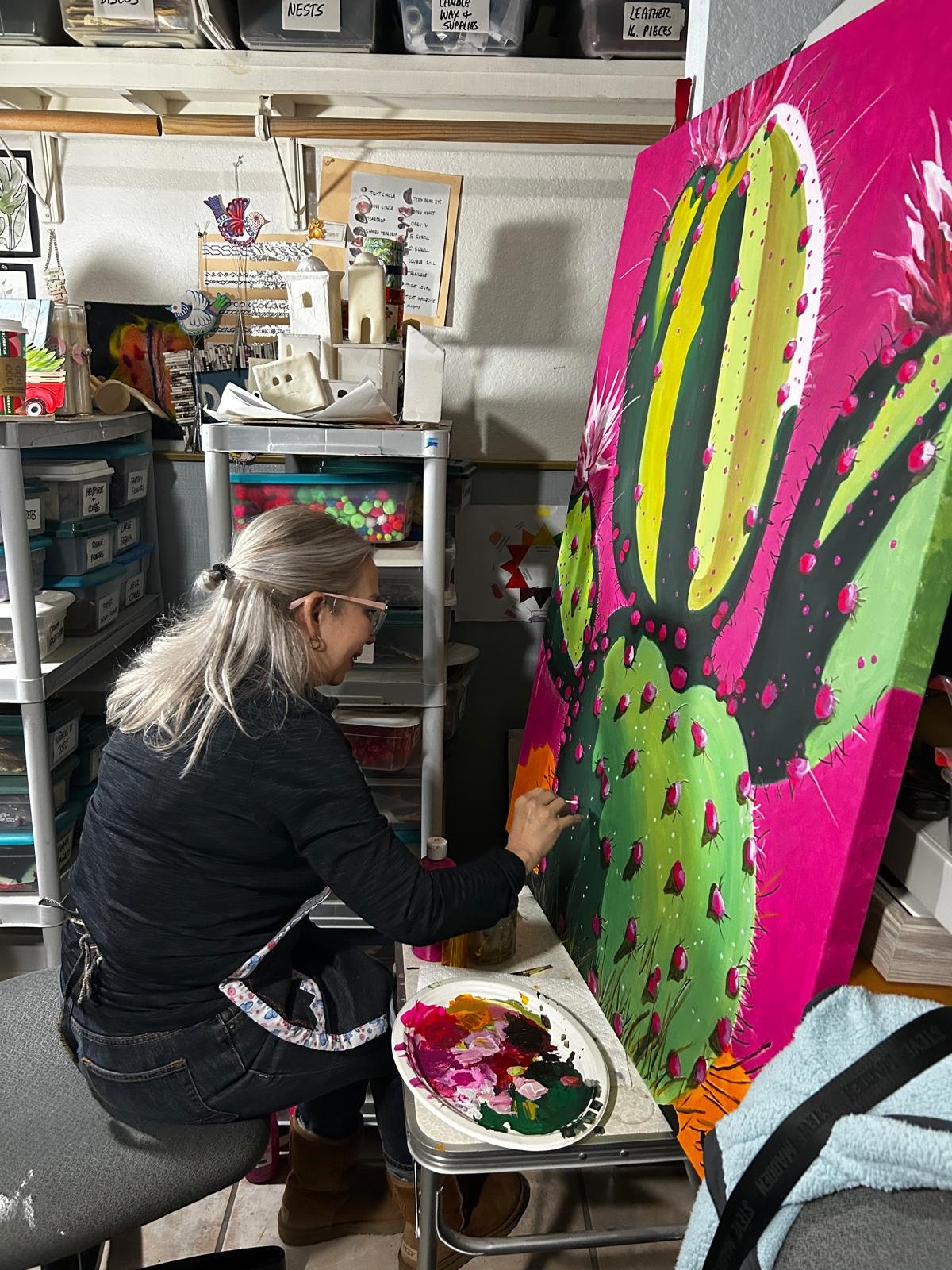 A woman with gray hair and glasses is sitting and painting a large colorful cactus on a canvas. She is in an art studio surrounded by shelves and storage bins filled with art supplies.