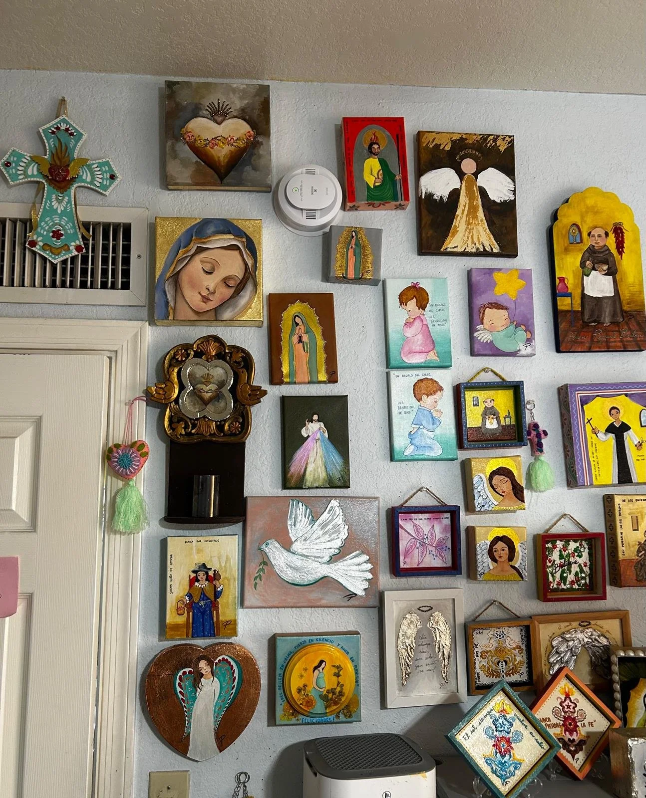 A wall decorated with numerous religious and angel-themed paintings and crafts, including crosses and a dove, in a home or church setting.