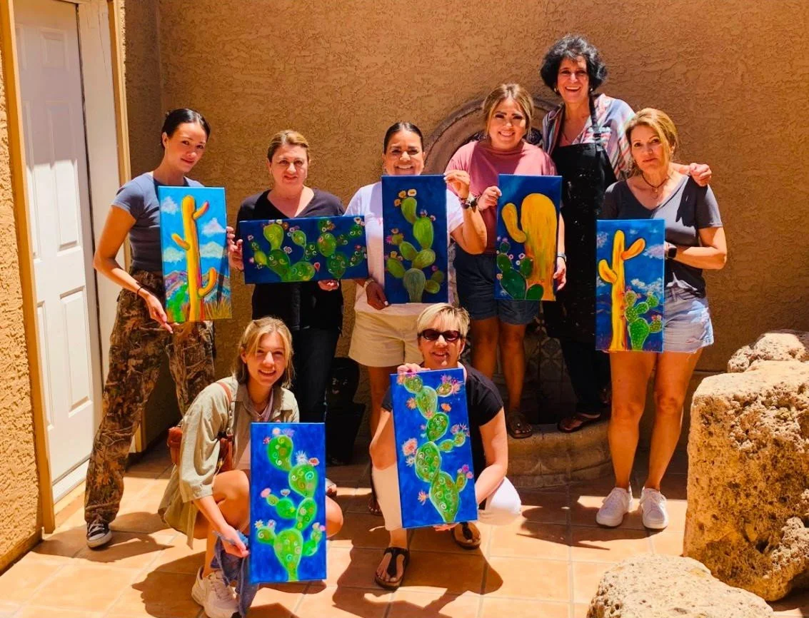 Group of ten women and girls holding cactus-themed paintings outdoors on a sunny day.