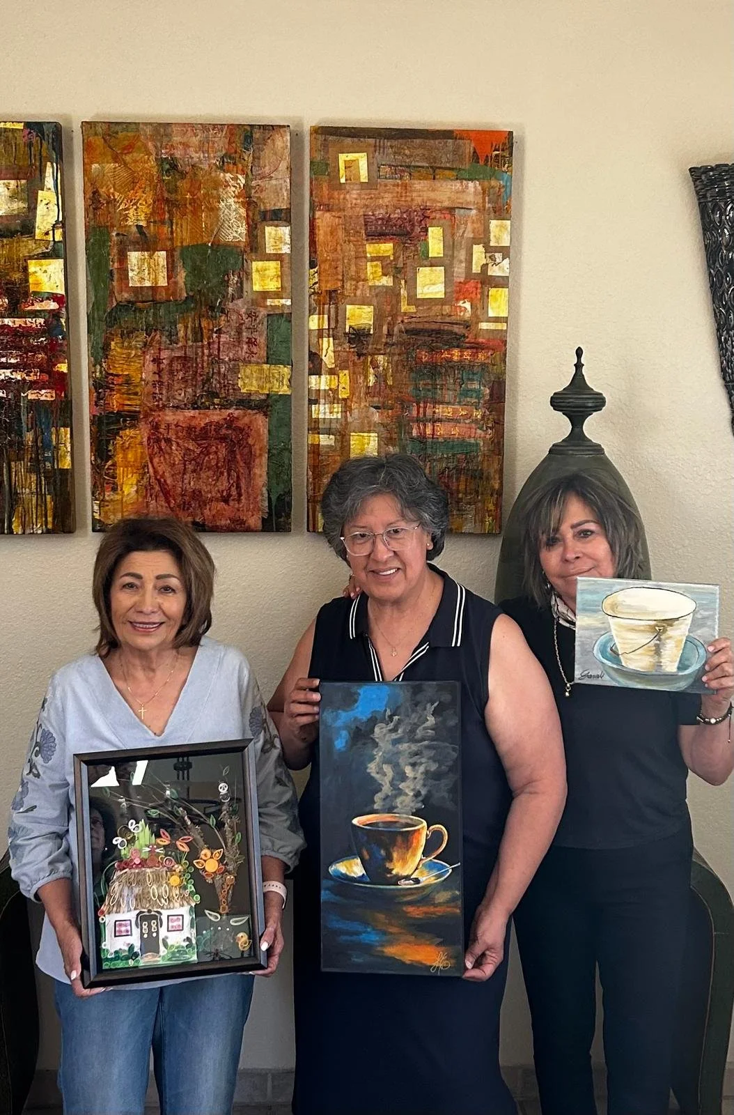 Three women standing in front of colorful abstract paintings, holding various pieces of artwork including a framed mosaic and paintings of coffee cups.