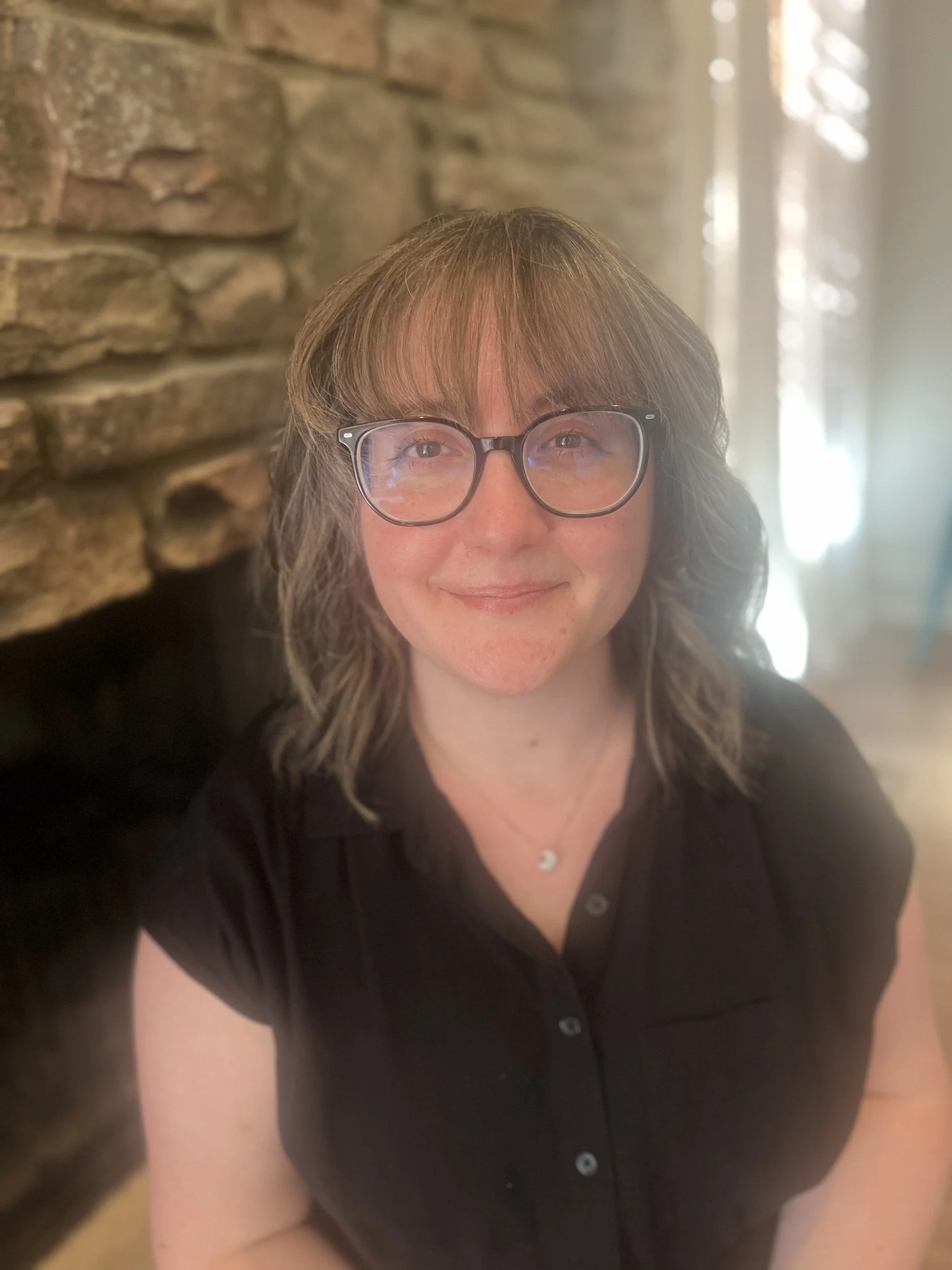 A woman with shoulder-length light brown hair, wearing glasses with a black frame, a black shirt, and a small crescent moon necklace. She is smiling and standing indoors near a stone wall with sunlight streaming in from behind.