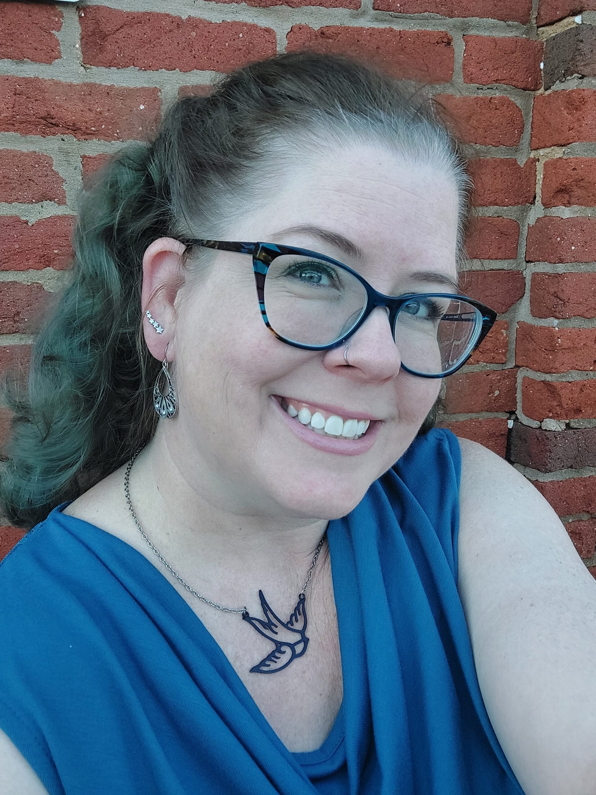 Close-up of a smiling woman with glasses, a tattoo of a bird on her collarbone, earrings, and a necklace, sitting against a brick wall.