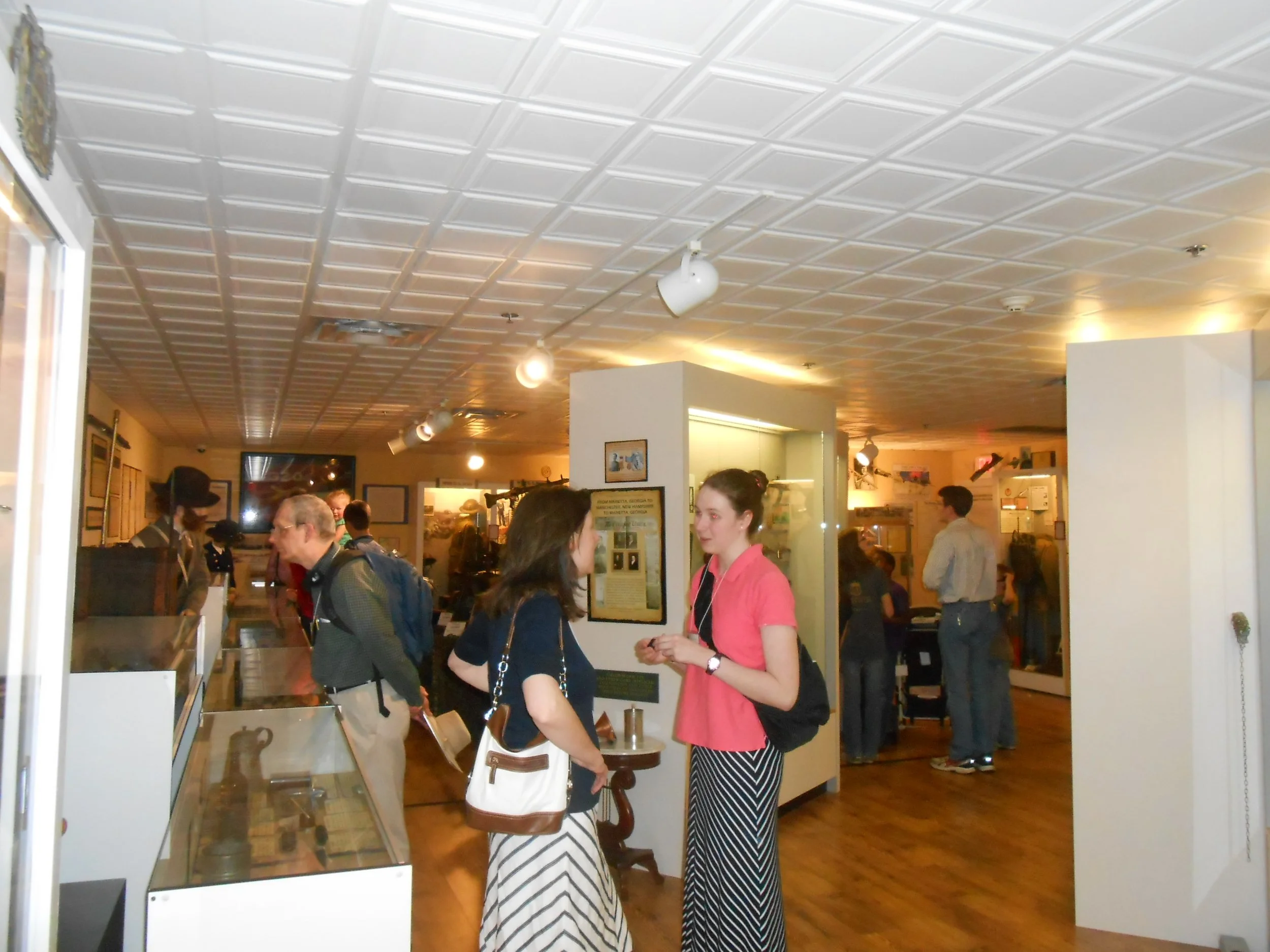 People inside a museum or historical exhibit, looking at display cases and information boards, with a decorative white ceiling and warm lighting.