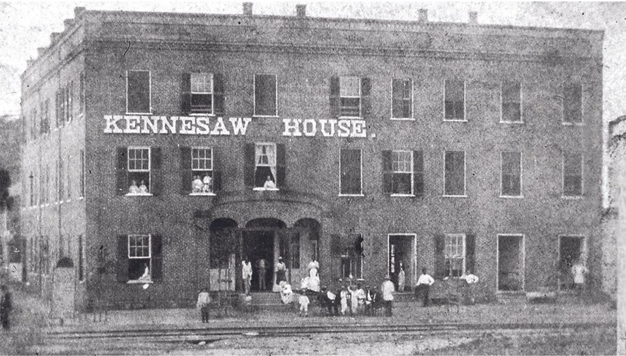 Historical black-and-white photo of a brick building labeled 'Kennesaw House' with people on the steps and around the entrance.