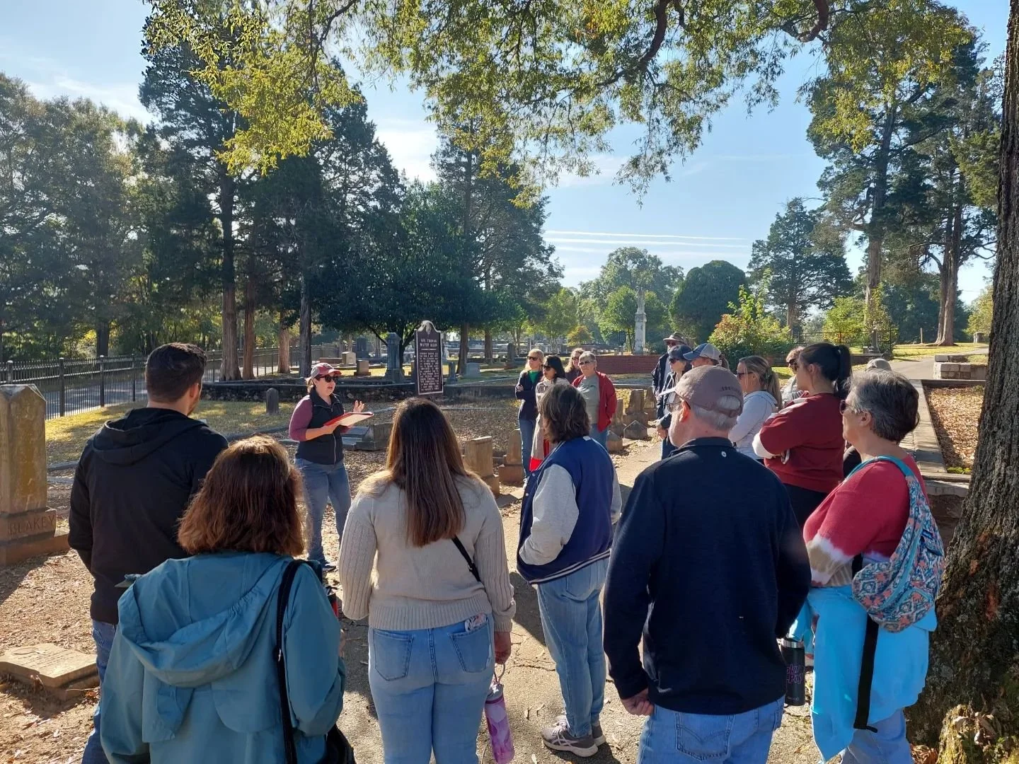 Group of people listening to a guide in a historic outdoor cemetery with trees and tombstones.