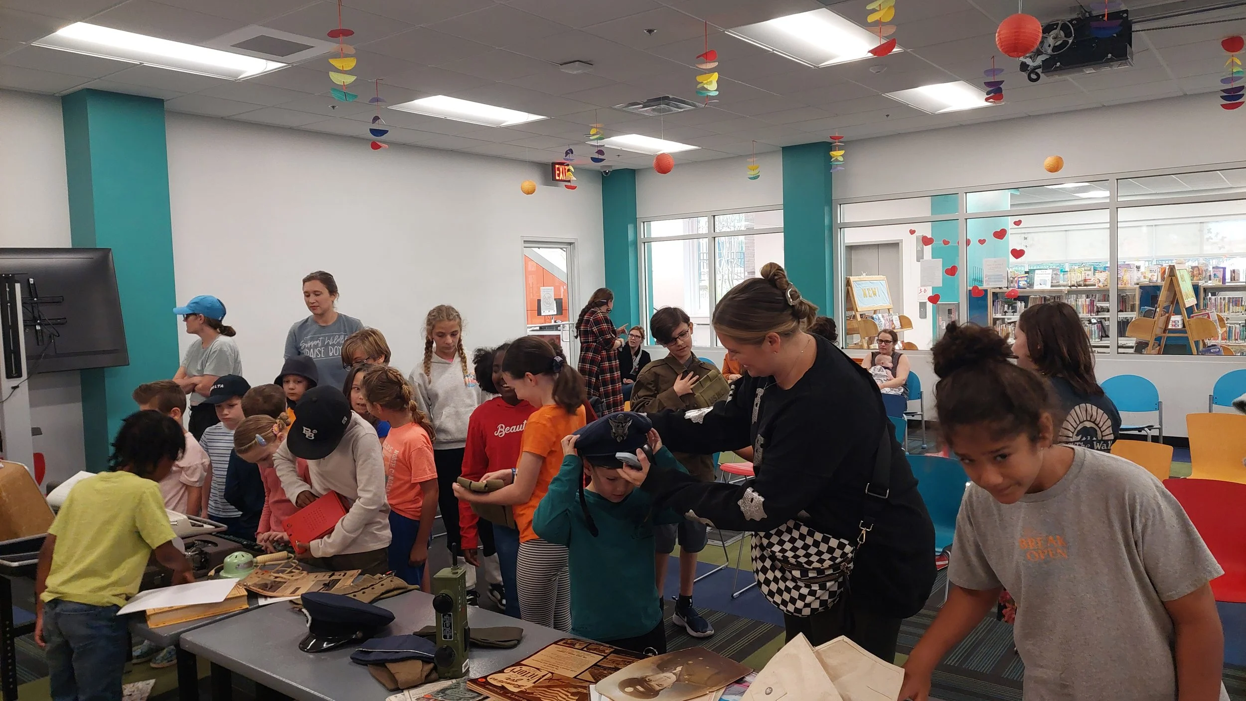 Children and adults gathered around tables in a colorful library, participating in a craft or activity event, with shelves of books and decorations hanging from the ceiling.