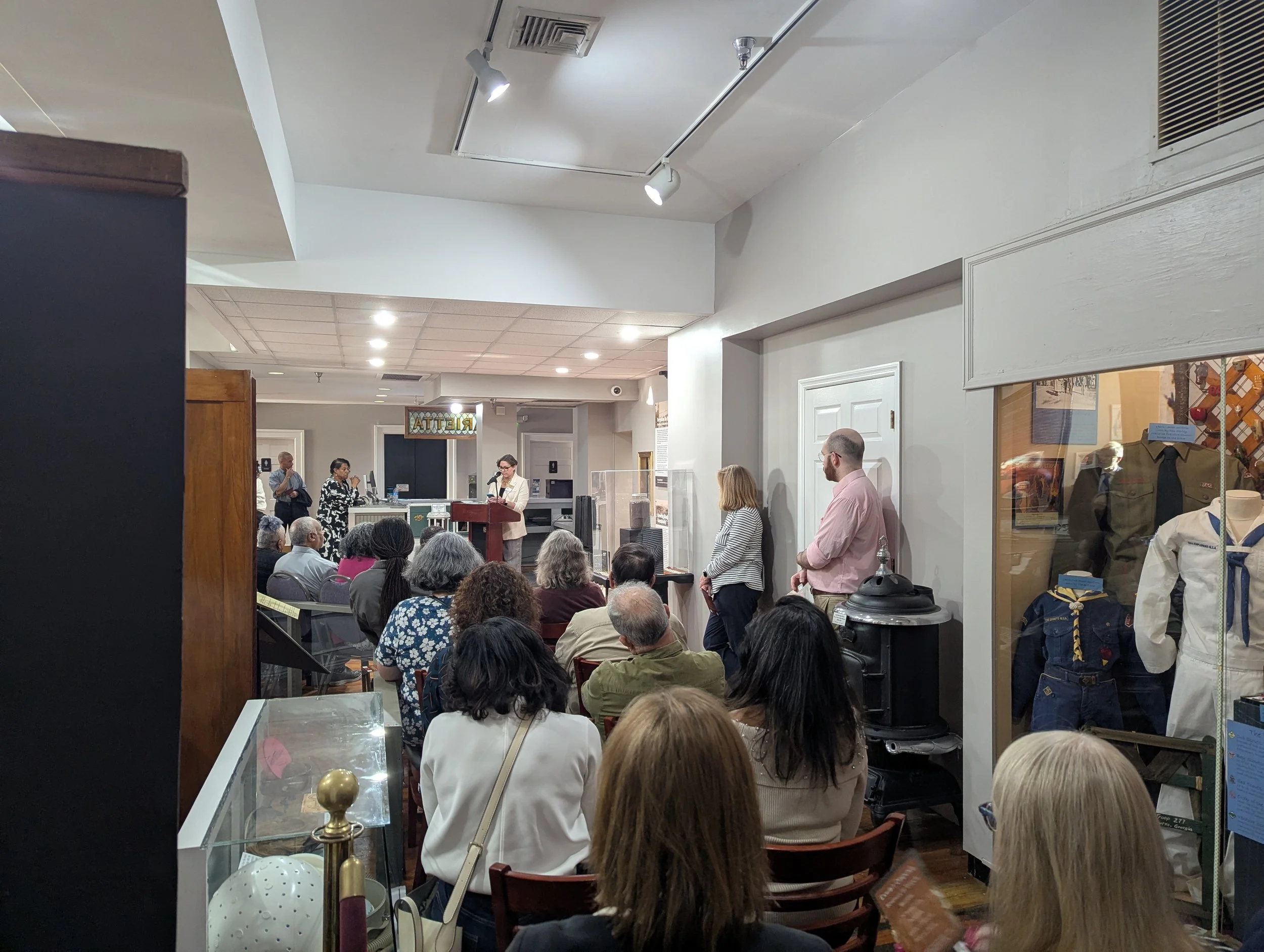 People gathered in a room attending an event or lecture, with some standing and some seated, and a woman speaking at a podium.
