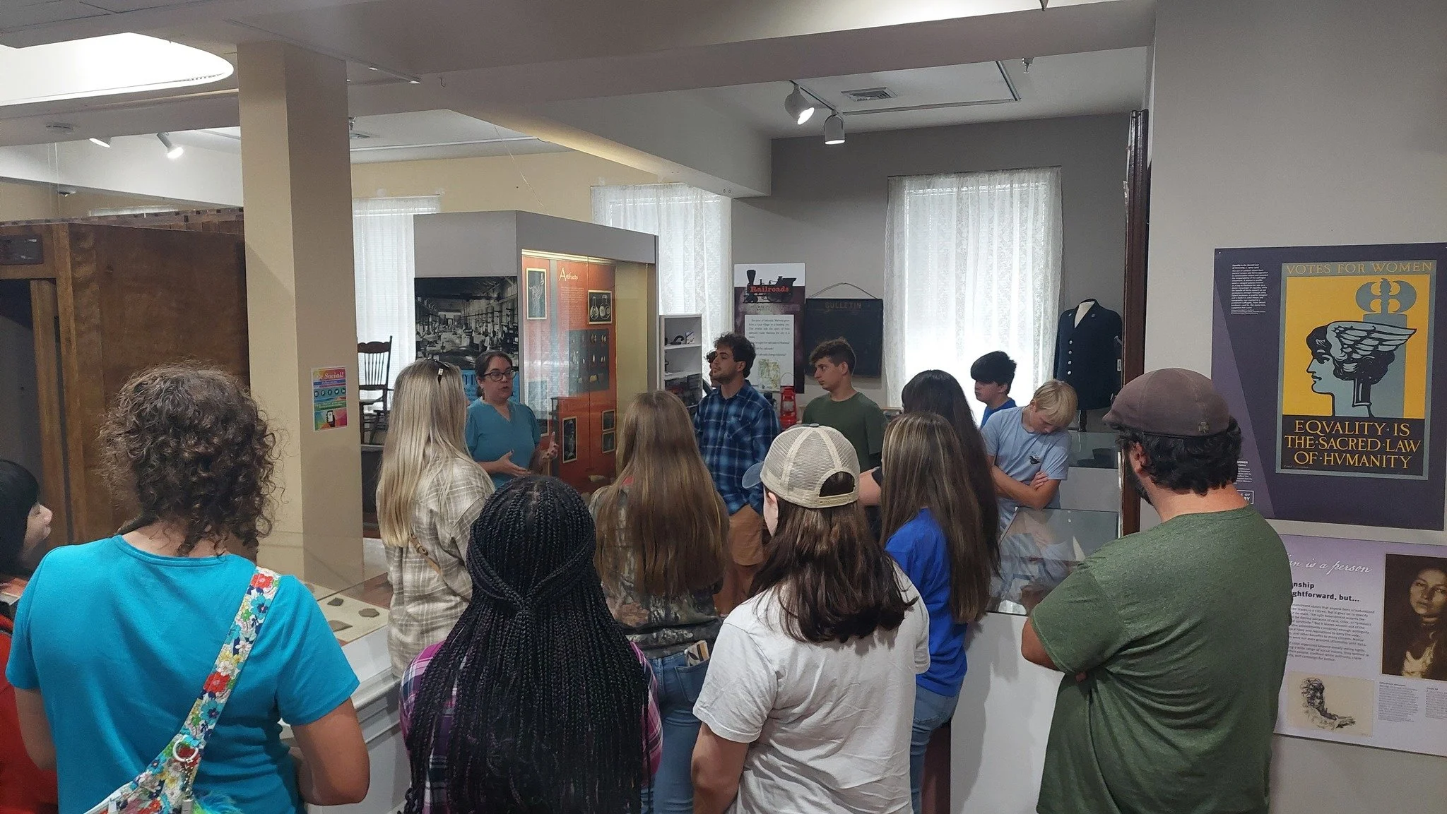 Group of people gathered around a woman giving a presentation in a museum or historical exhibit, with posters and information displays on the walls.