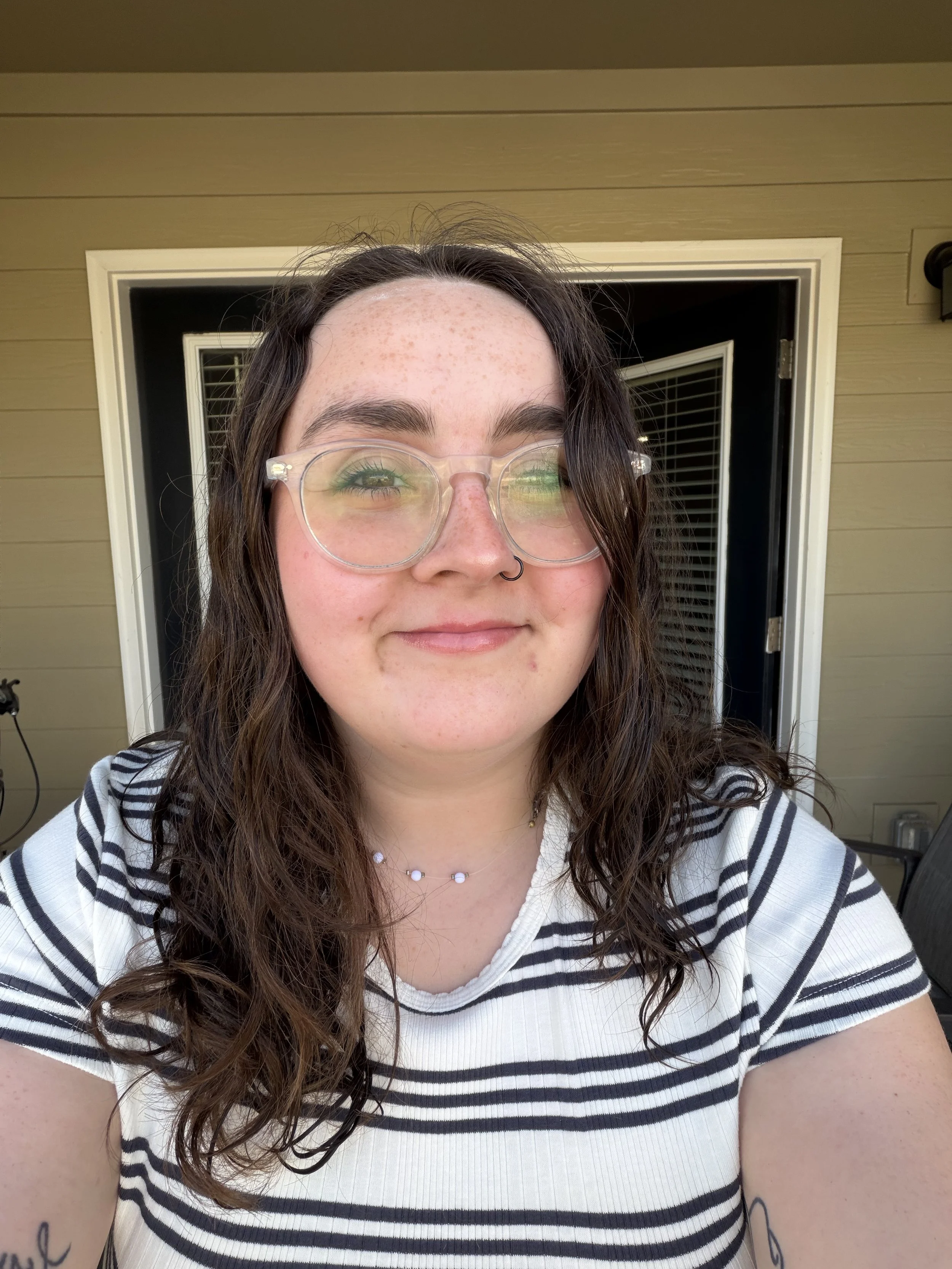 A young woman with long, wavy brown hair, wearing clear glasses with a slight green reflection, a black nose ring, a beaded necklace, and a white shirt with black horizontal stripes, is smiling outdoors in front of a beige house with white trim and a door with blinds.