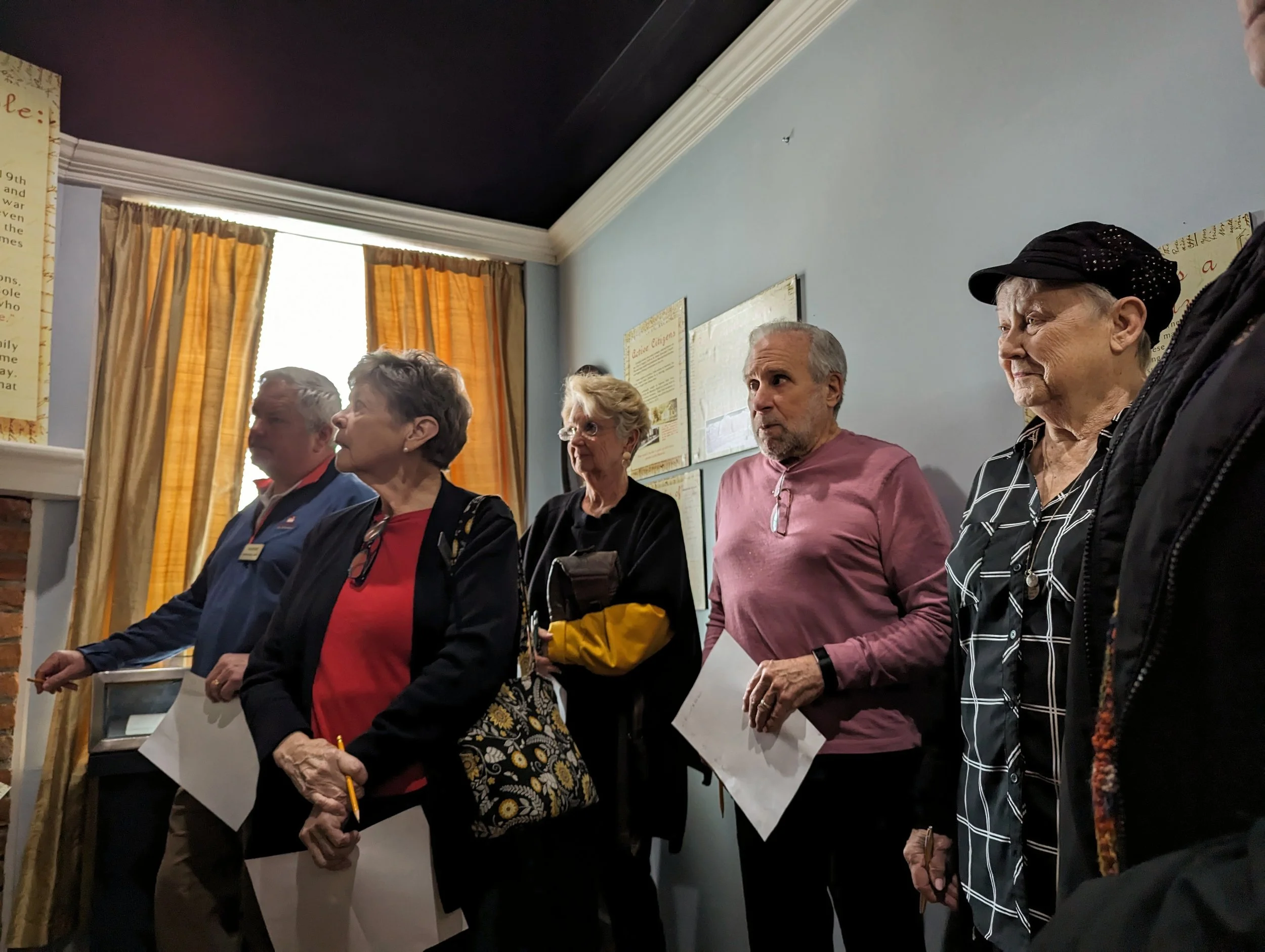 A group of elderly people standing closely together in a room with cream curtains and framed posters on the wall.