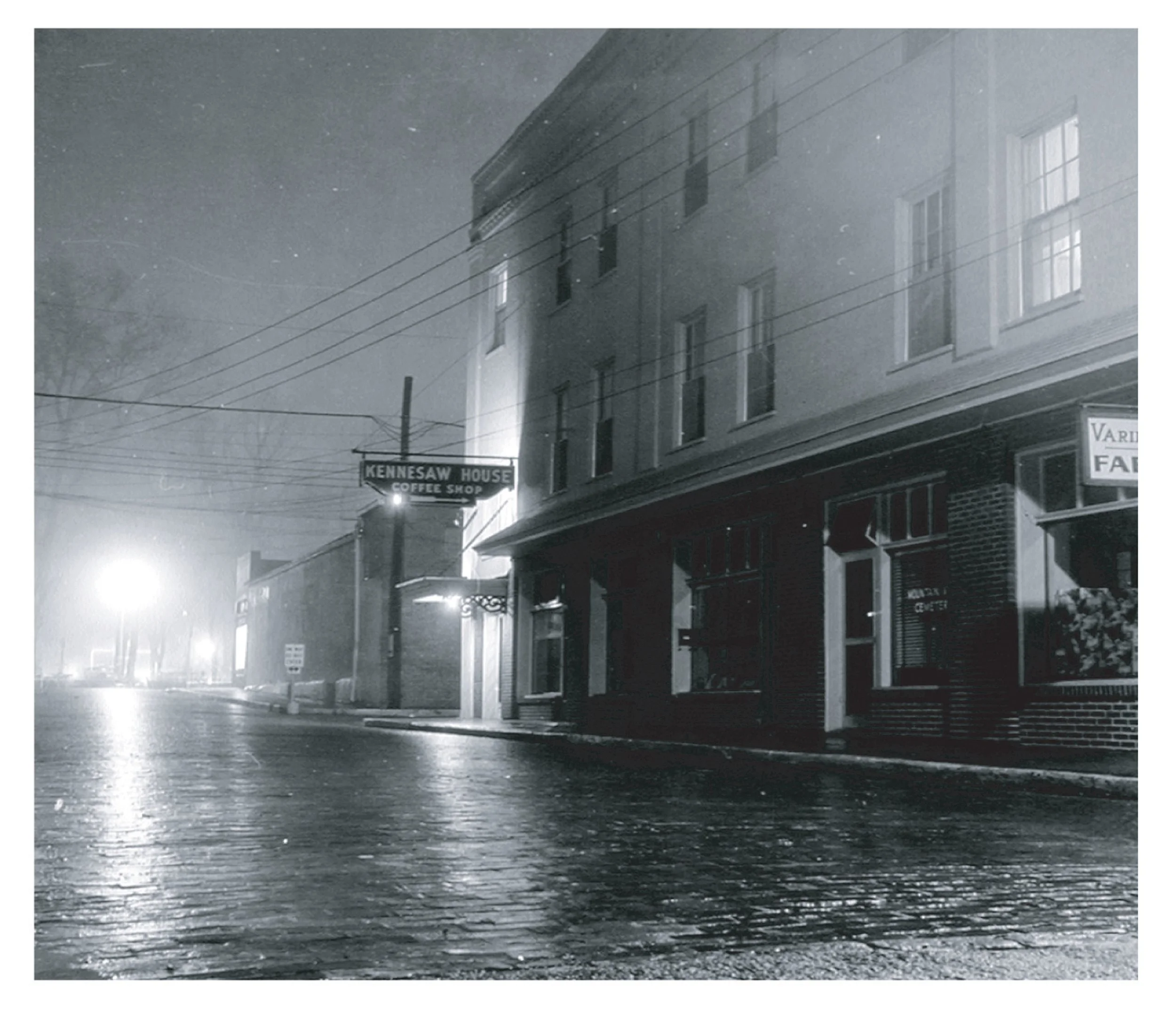 Night scene of a city street during a rainstorm, with reflections on the wet pavement, illuminated by streetlights and building lights, and a sign for Kennesaw House Coffee Shop.