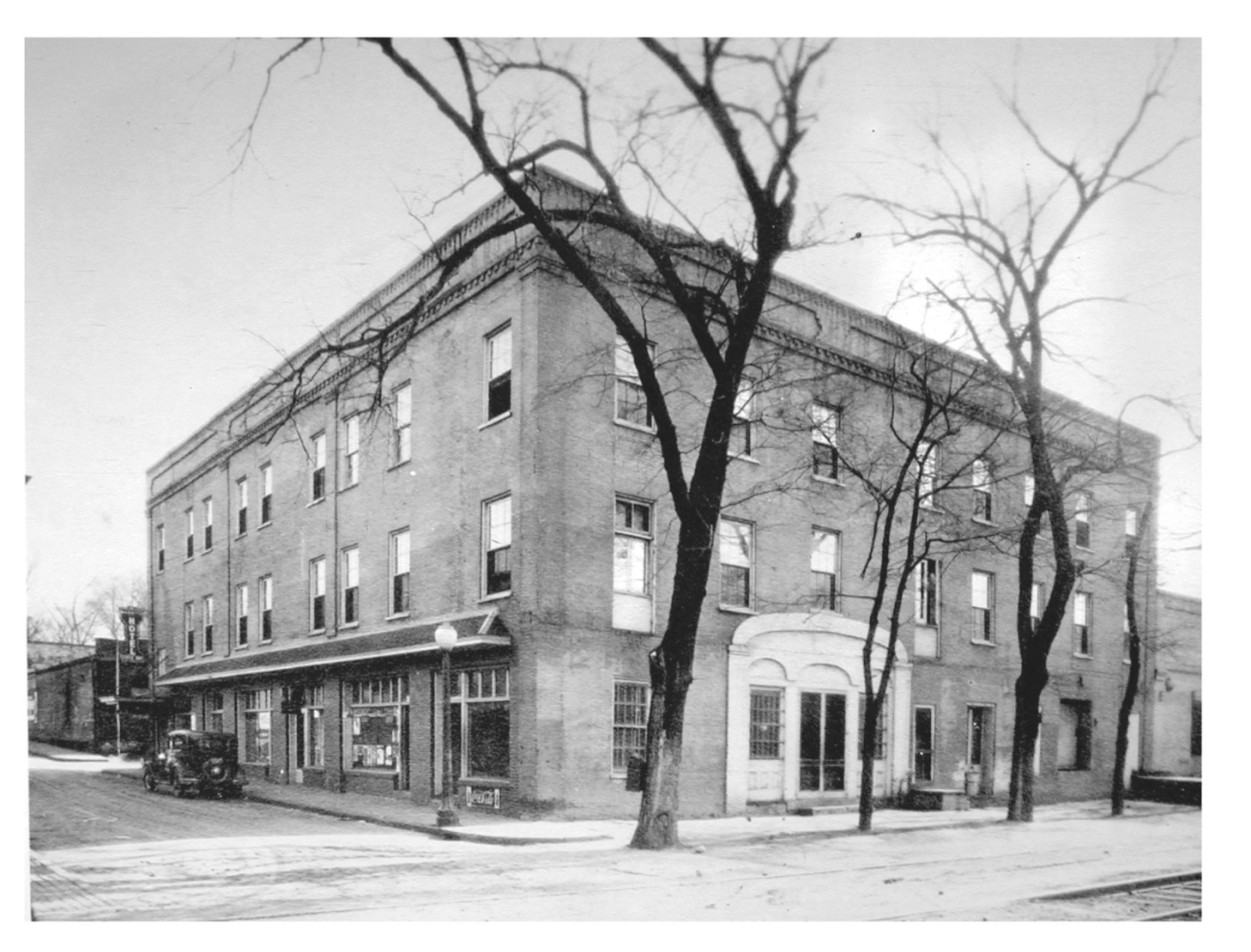 Black and white photo of a building with multiple windows, trees with bare branches in the foreground, and a small vehicle parked in front.