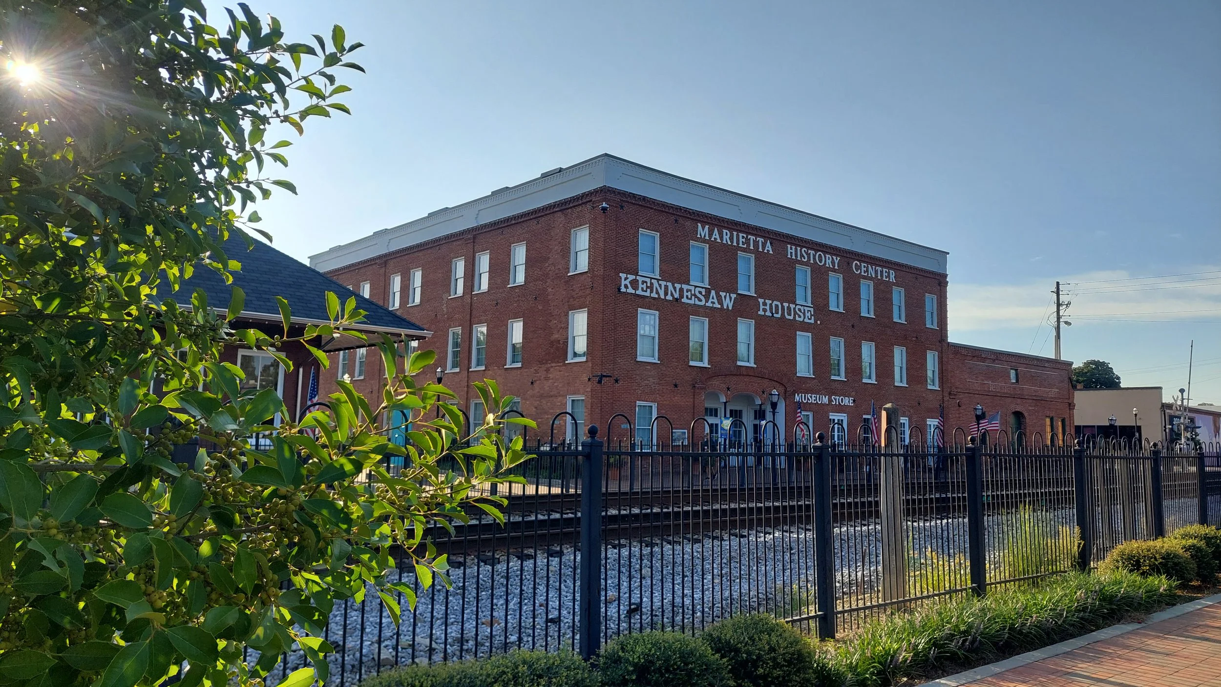 Historic brick building labeled as Marietta History Center and Kennesaw House, beside a railway track, with trees, a fence, and a sidewalk in the foreground.