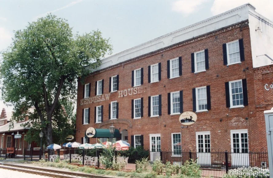 Historic brick building named 'Kenosha House' with black shutters, outdoor seating with umbrellas, a large tree, and decorative signs.