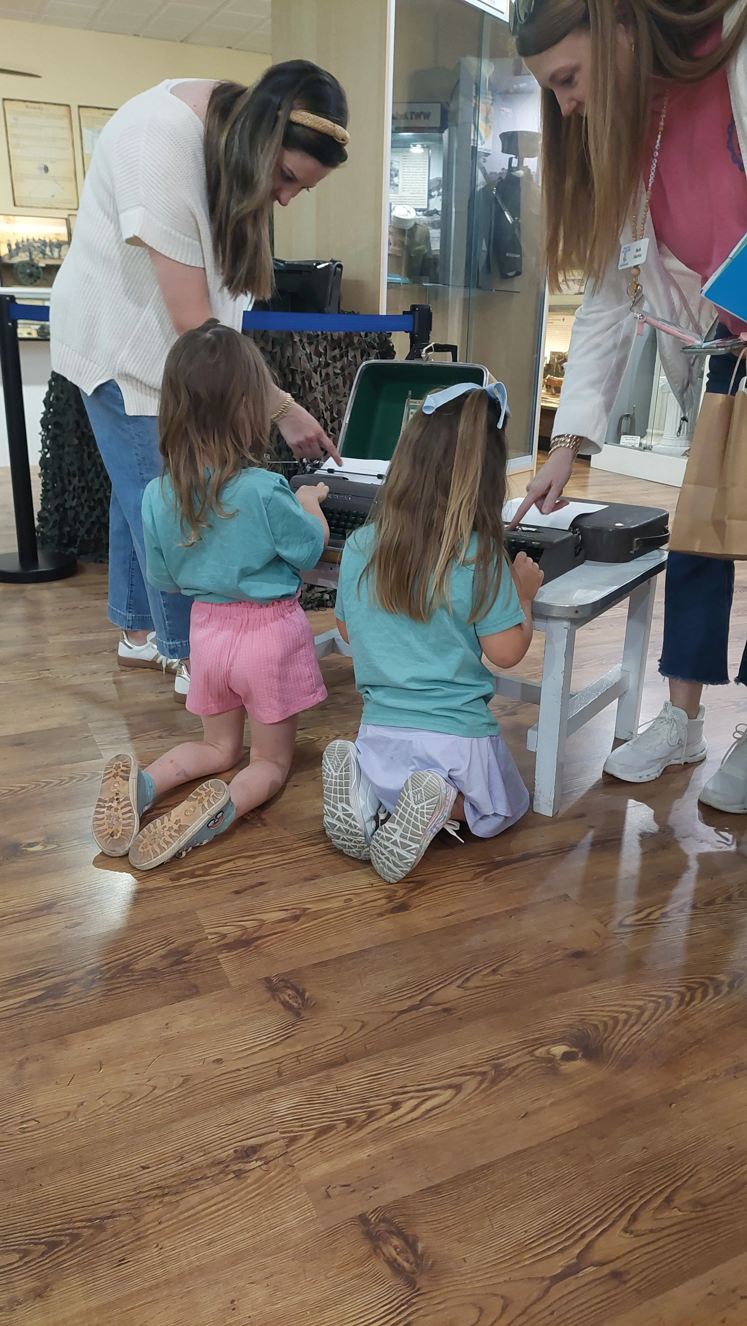 Two young girls kneeling on the hardwood floor, working on a vintage typewriter, with two women standing beside them, pointing at the typewriter, in a museum or exhibit setting.