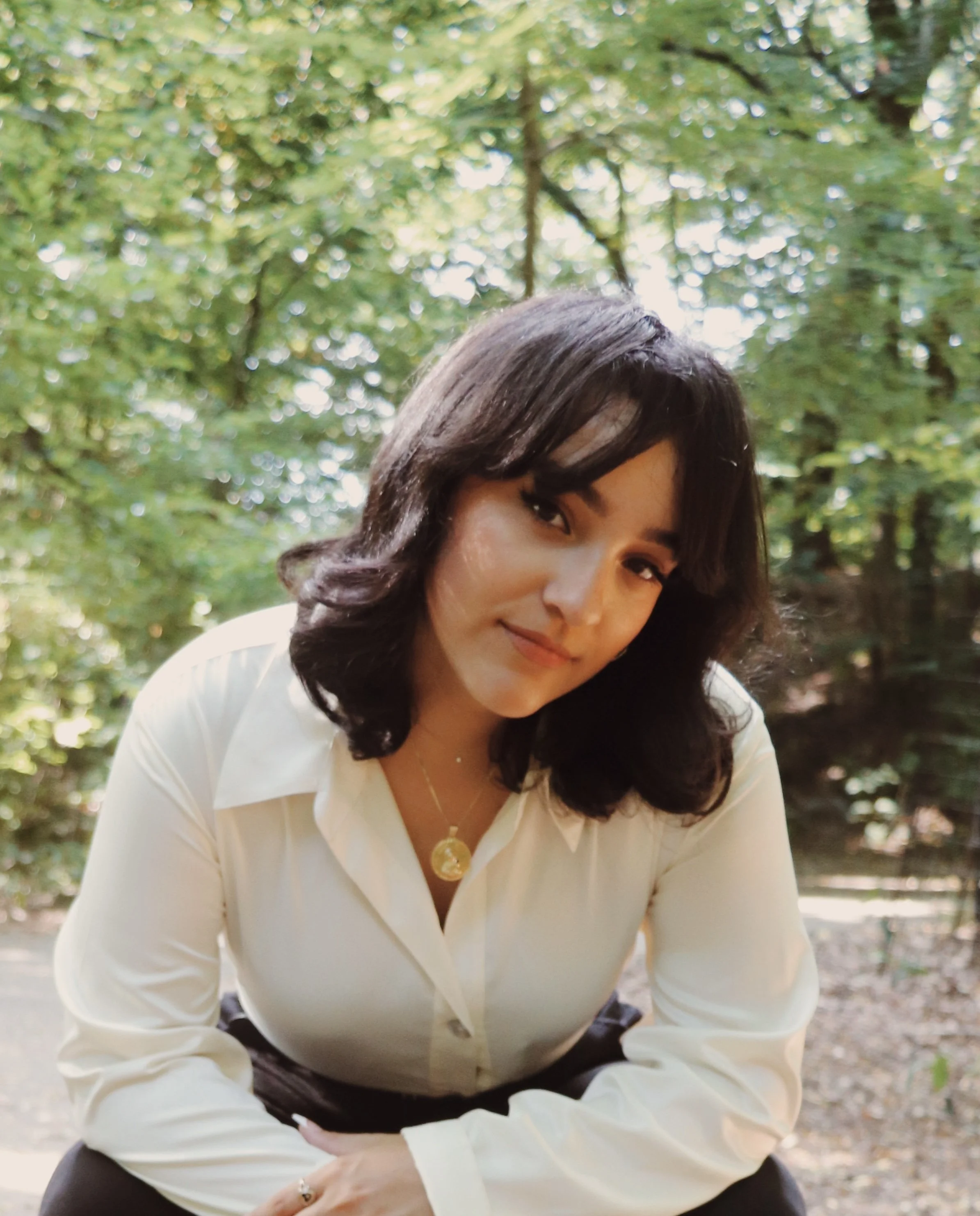 A woman with dark, shoulder-length hair and a friendly expression, wearing a cream-colored blouse and gold necklace, outdoors in a lush, green wooded area.