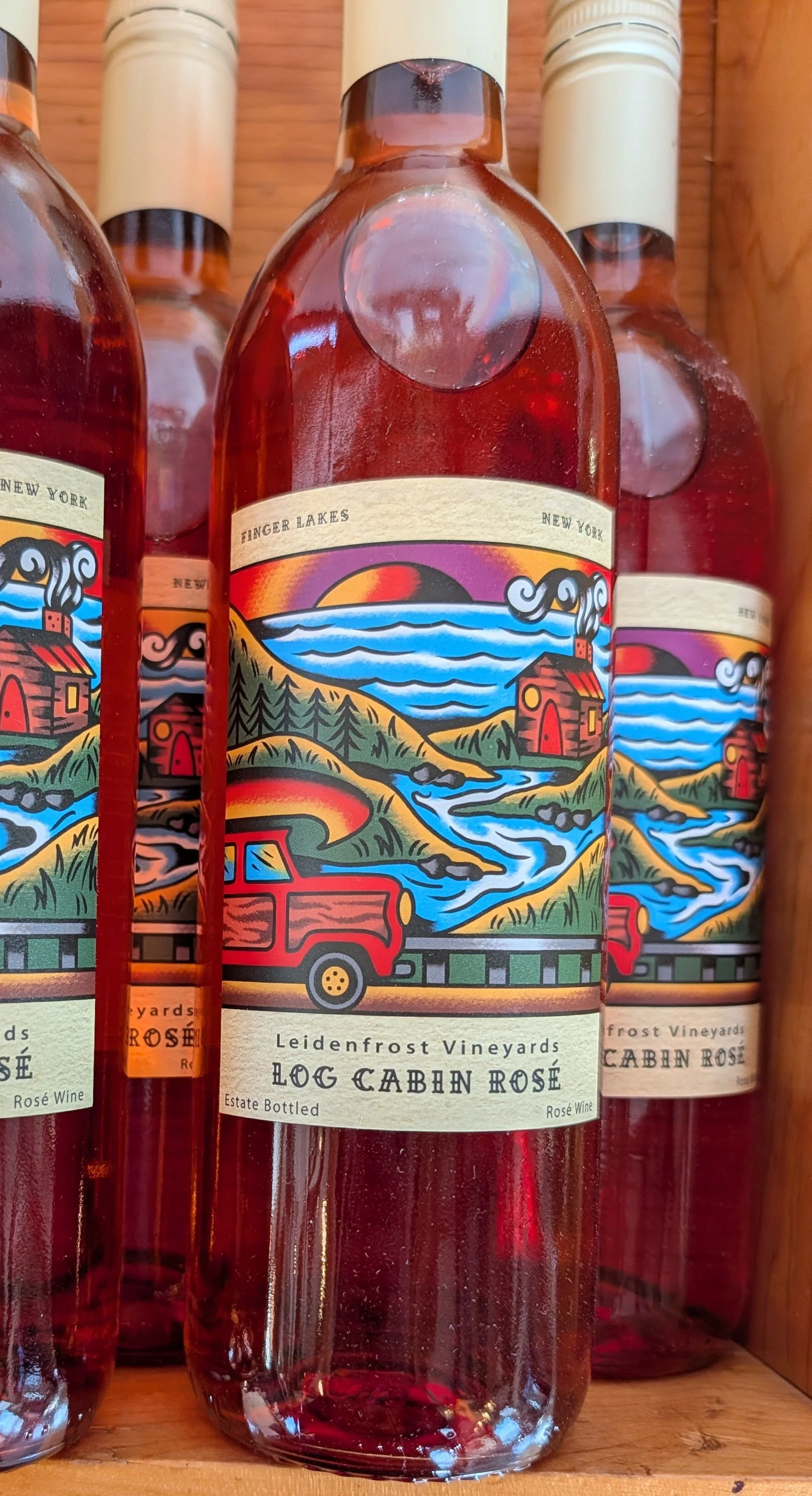 A glass bottle of Log Cabin Rosé wine from Finger Lakes, New York, with a colorful landscape label featuring a red truck, a river, hills, trees, and a sunset.