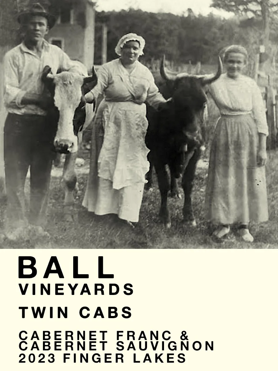 Black and white historic photo of three people standing outdoors with two cows. The man on the left holds a cow's head, the woman in the middle holds the cow's horns, and the woman on the right stands beside the second cow. There are trees and a house in the background.