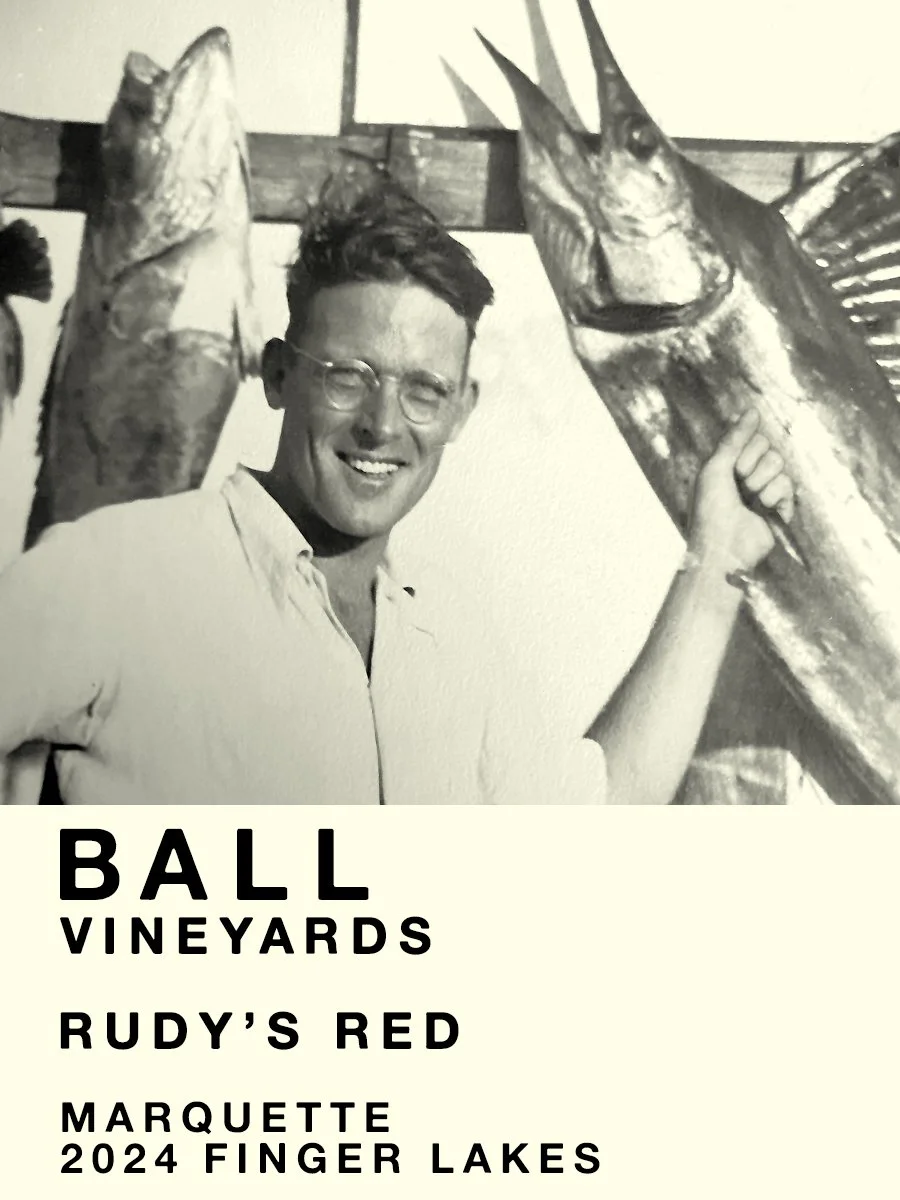 A black-and-white photo of a smiling man wearing glasses and a white shirt, holding a large fish. Behind him, there are more fish hanging. At the bottom of the image, there is text that reads: "Ball Vineyards Rudy's Red Marquette 2024 Finger Lakes".