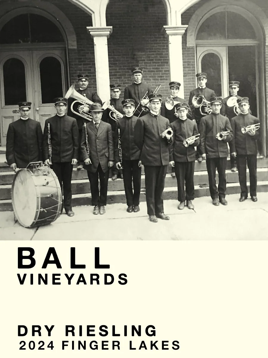 Black and white photo of a marching band with young men in uniform holding musical instruments, standing on steps in front of a brick building with arched windows and columns, labeled as BALL VINEYARDS DRY RIESLING 2024 FINGER LAKES.
