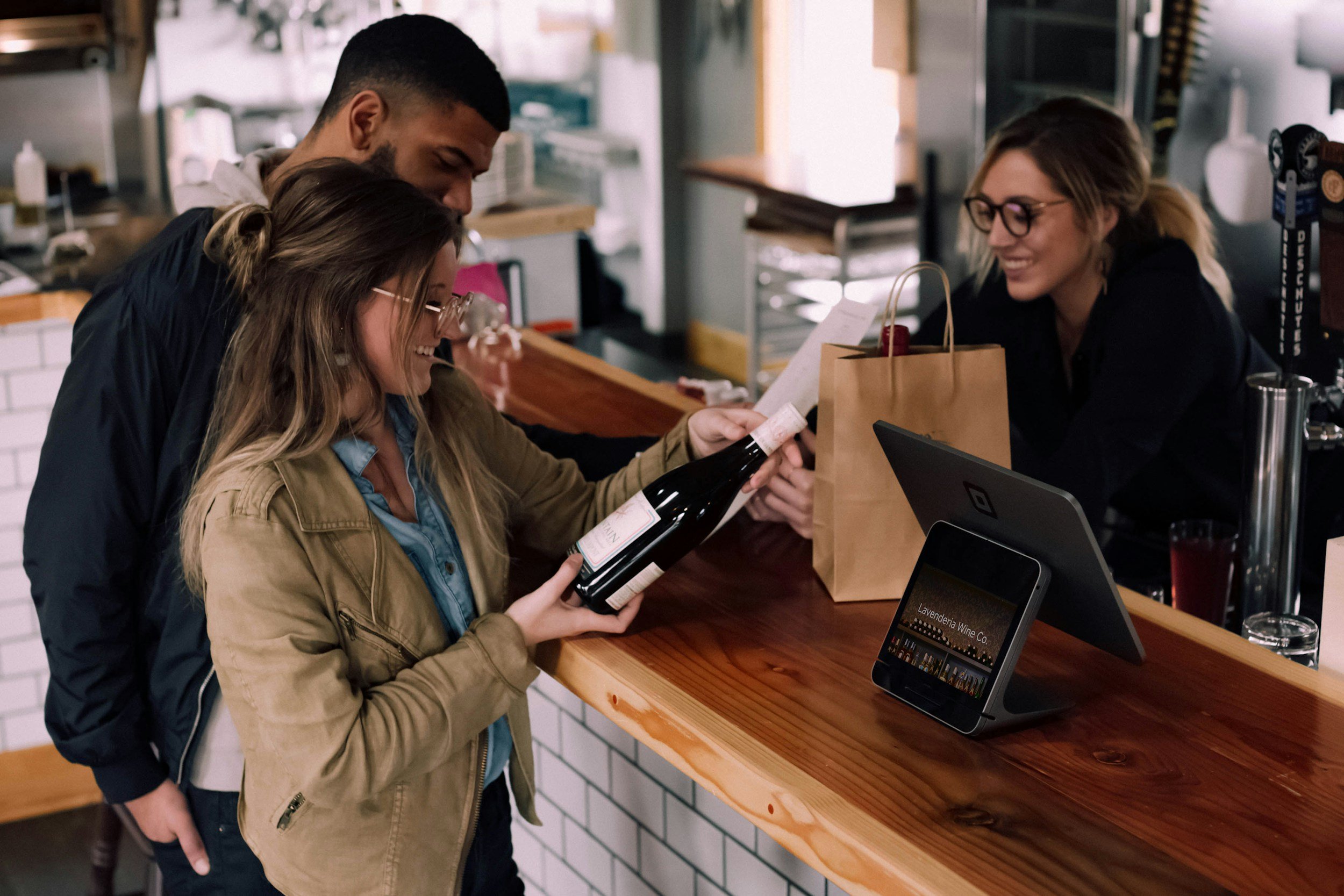 Two customers at a bar counter, a man and a woman, are selecting a bottle of wine, while the bartender, a woman with glasses, smiles at them. The counter has a tablet device, a paper bag, and drinks.