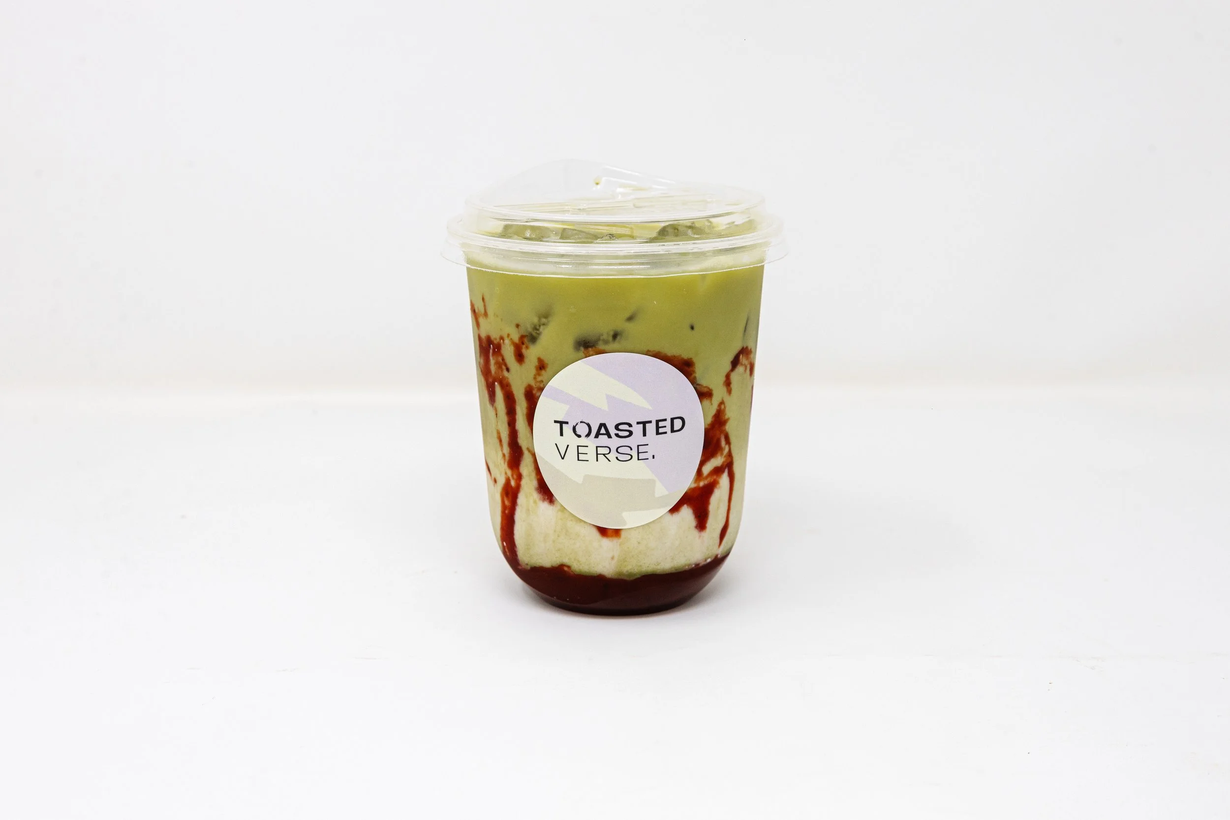 A clear plastic cup of green iced matcha latte with caramel and red syrup swirls, labeled 'Toasted Verse,' against a plain white background.