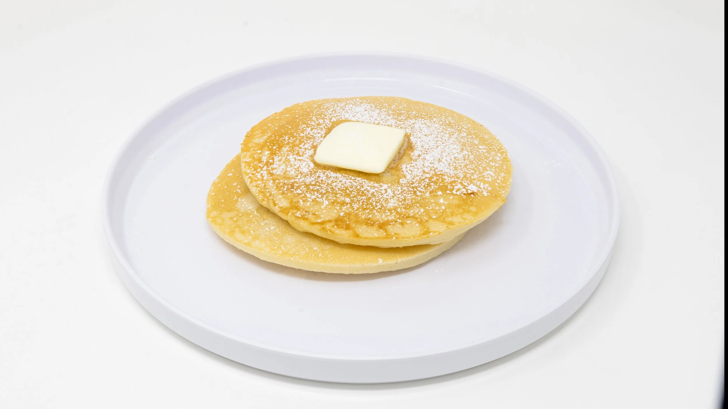 Two pancakes with a pat of butter on top, dusted with powdered sugar, on a white plate against a white background.