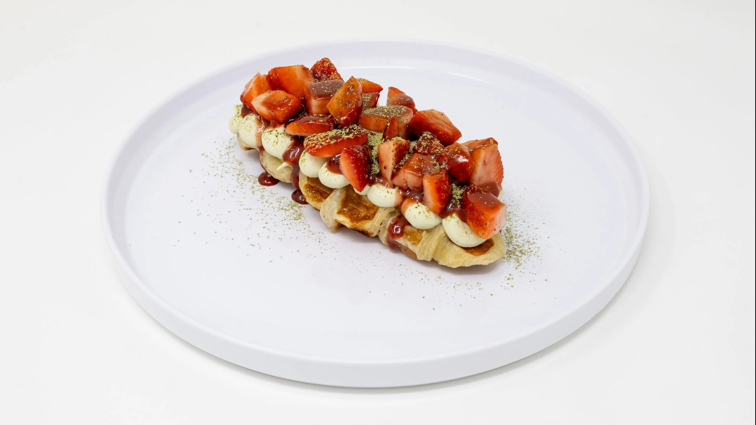 A plated dessert with sliced bananas topped with strawberries, whipped cream, and caramel sauce, garnished with green powder, served on a white circular plate.