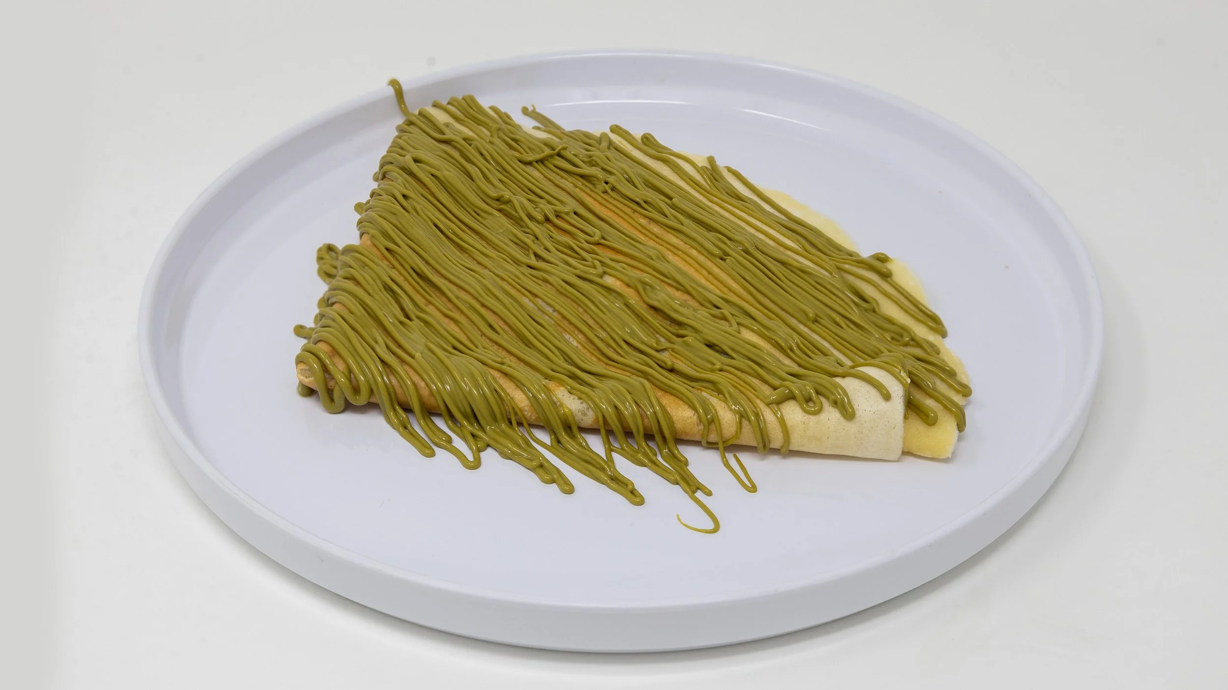 Unrolled crepe on a white plate topped with green matcha-flavored icing drizzled in a zigzag pattern.