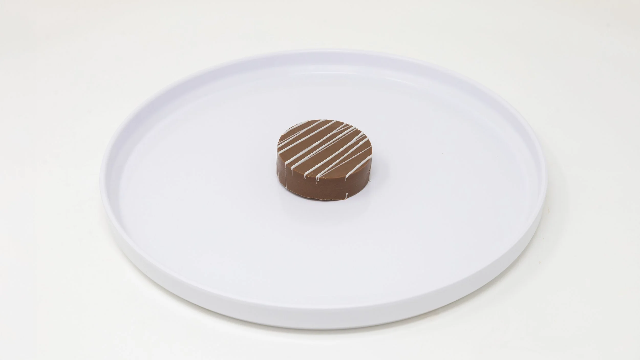 A round chocolate dessert with white drizzle on top, placed on a white circular tray.