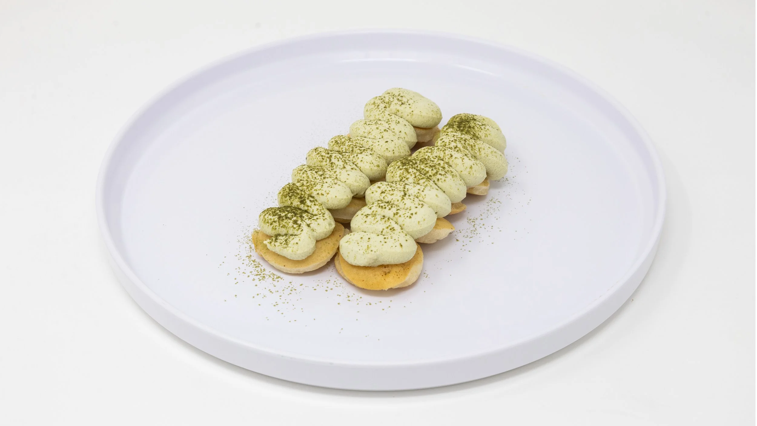 A white oval plate with twenty small cookies topped with white cream and sprinkled with green powder.