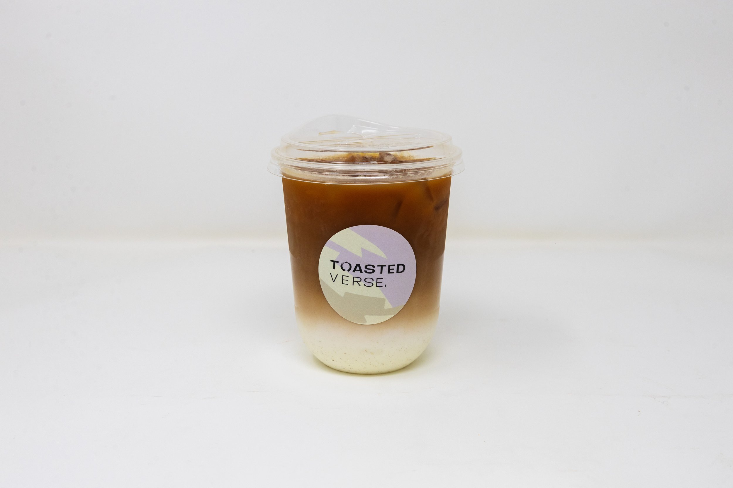 A plastic cup filled with iced coffee layered with milk, with a clear lid and a sticker that says 'Toasted Verse.'