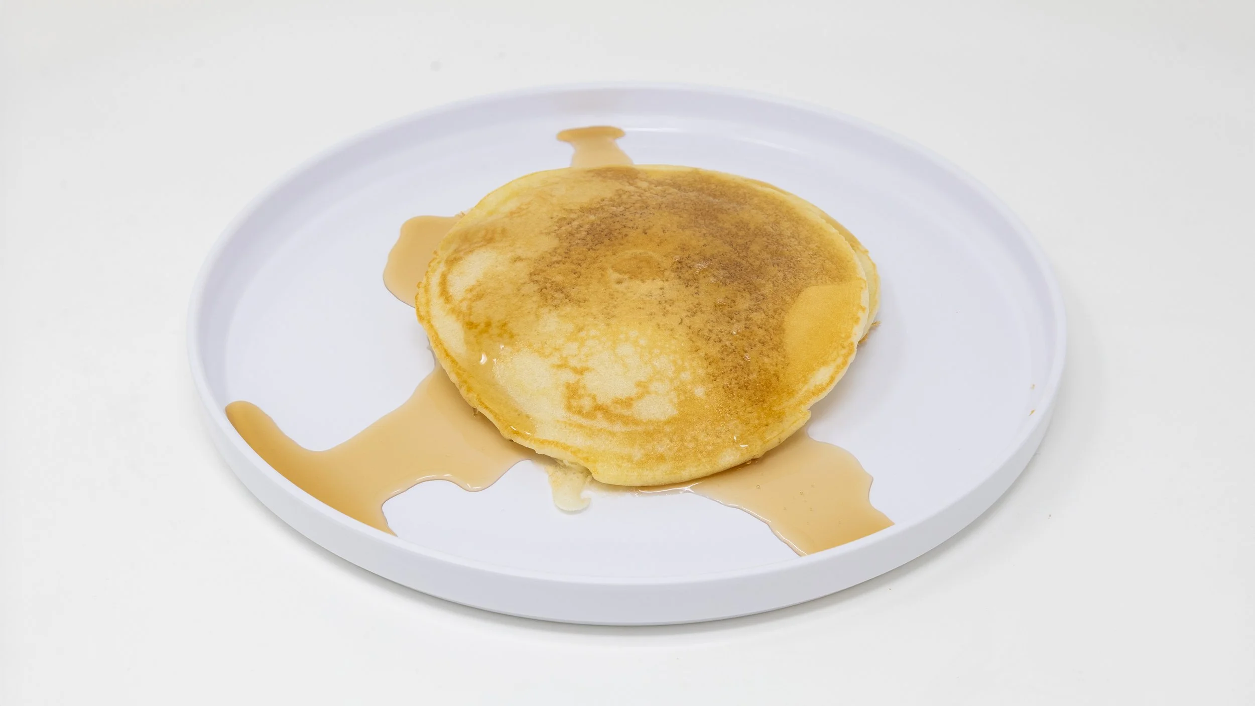 A stack of pancakes on a white plate with syrup on top and around the pancakes, against a white background.